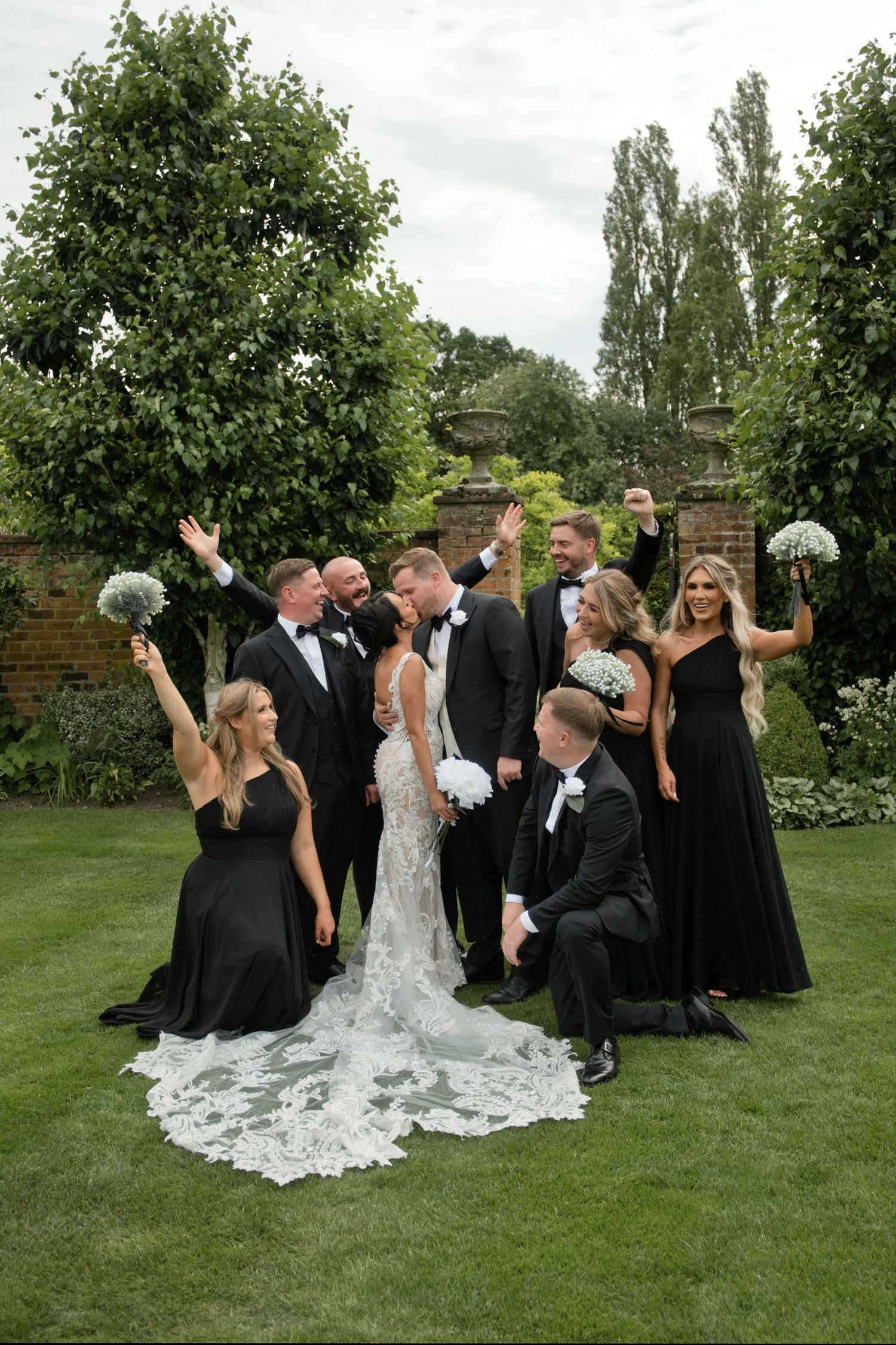Wedding party celebrates outdoors in garden, bridesmaids in black gowns and groomsmen in black tuxedos surround kissing couple.