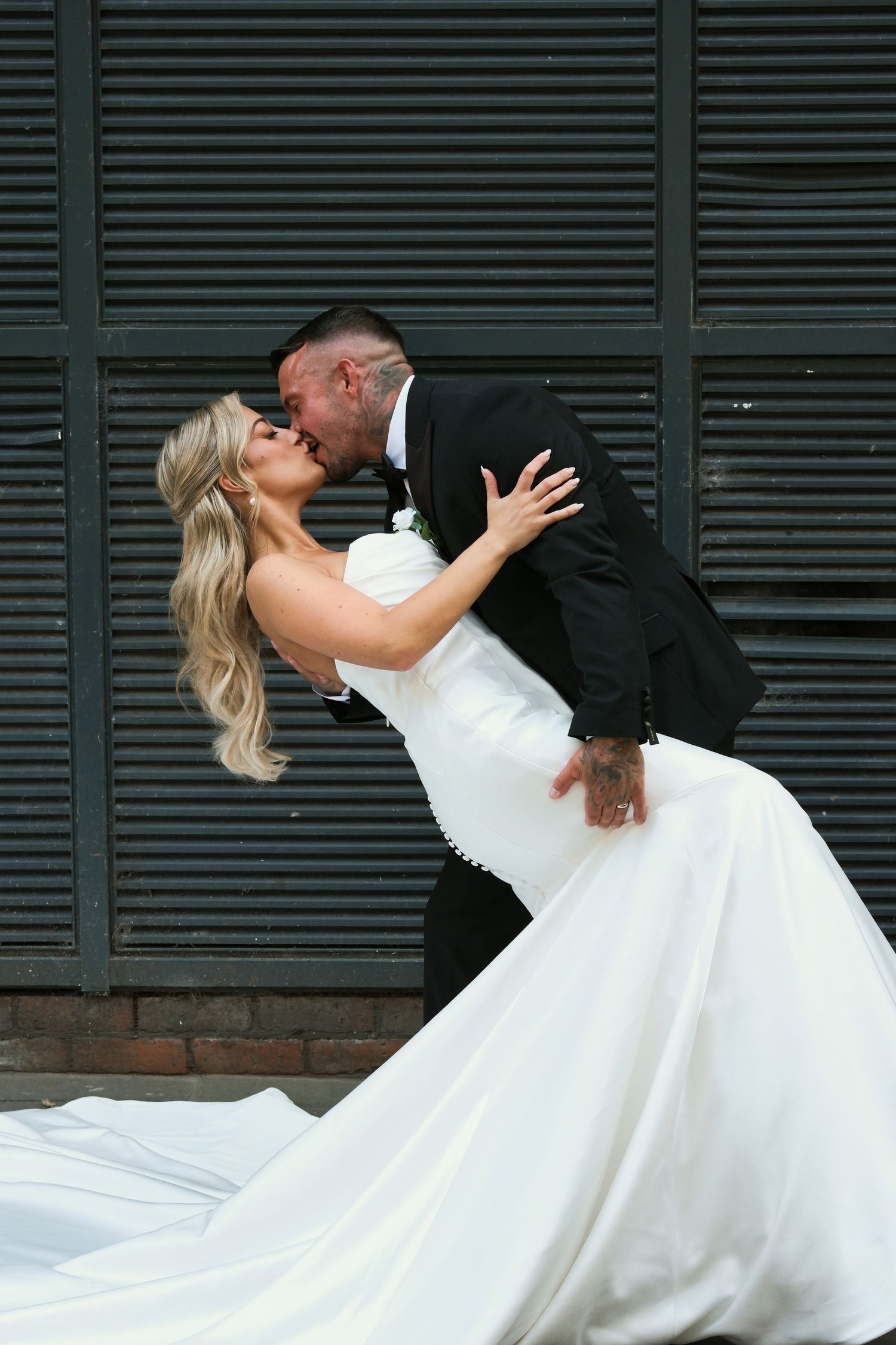 Groom dips bride in white mermaid gown for a romantic kiss against dark shuttered backdrop.