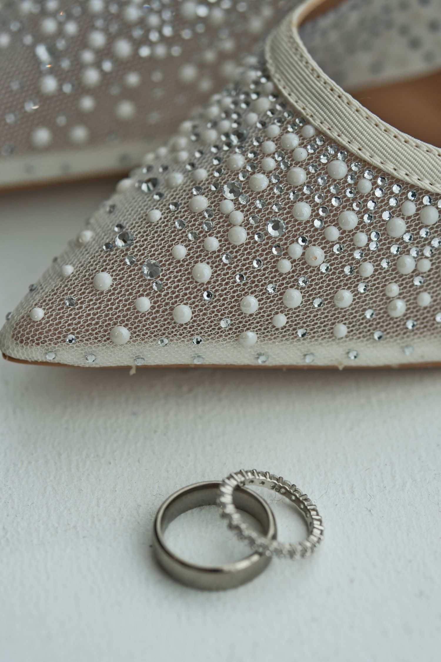 Close-up of pearl and crystal studded wedding heels with two silver wedding bands on white surface.