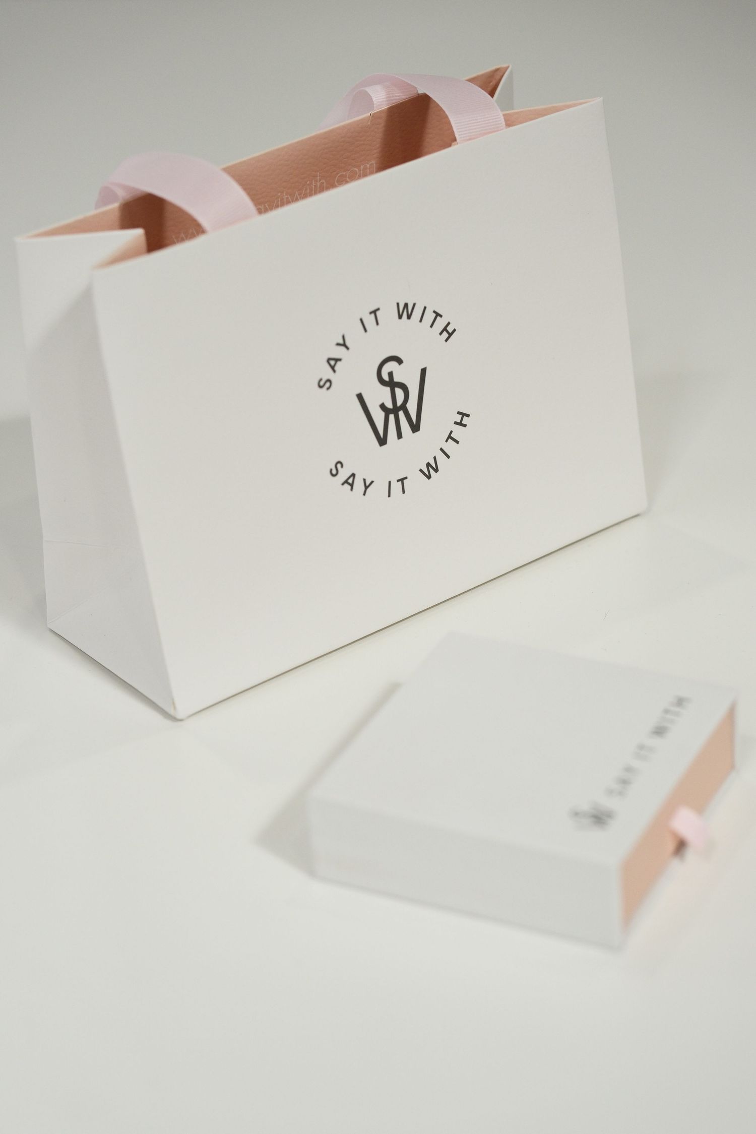 Elegant white gift bag and box with pink ribbon handles featuring Say It With brand logo on neutral background.