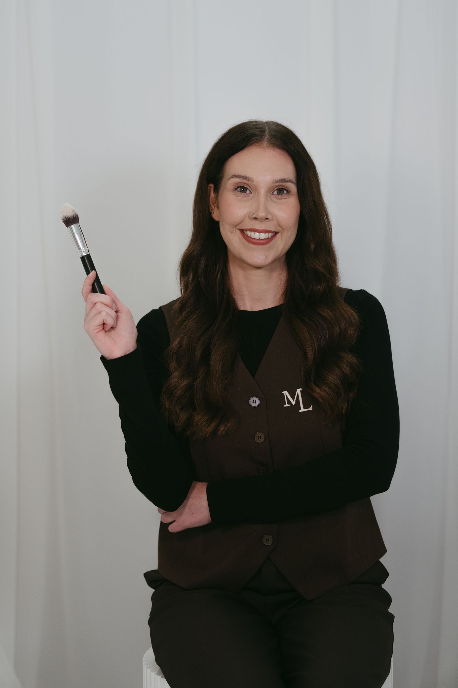 Smiling makeup artist holding a brush, wearing a brown monogrammed vest over a black top against a white background.