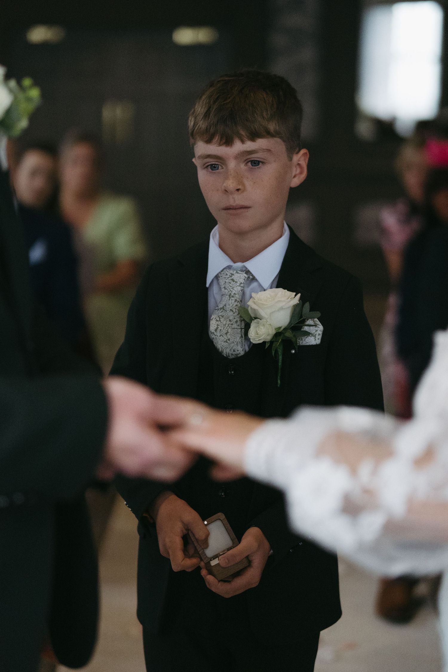 Young ring bearer in black suit with white rose boutonniere holds ring box at wedding ceremony.
