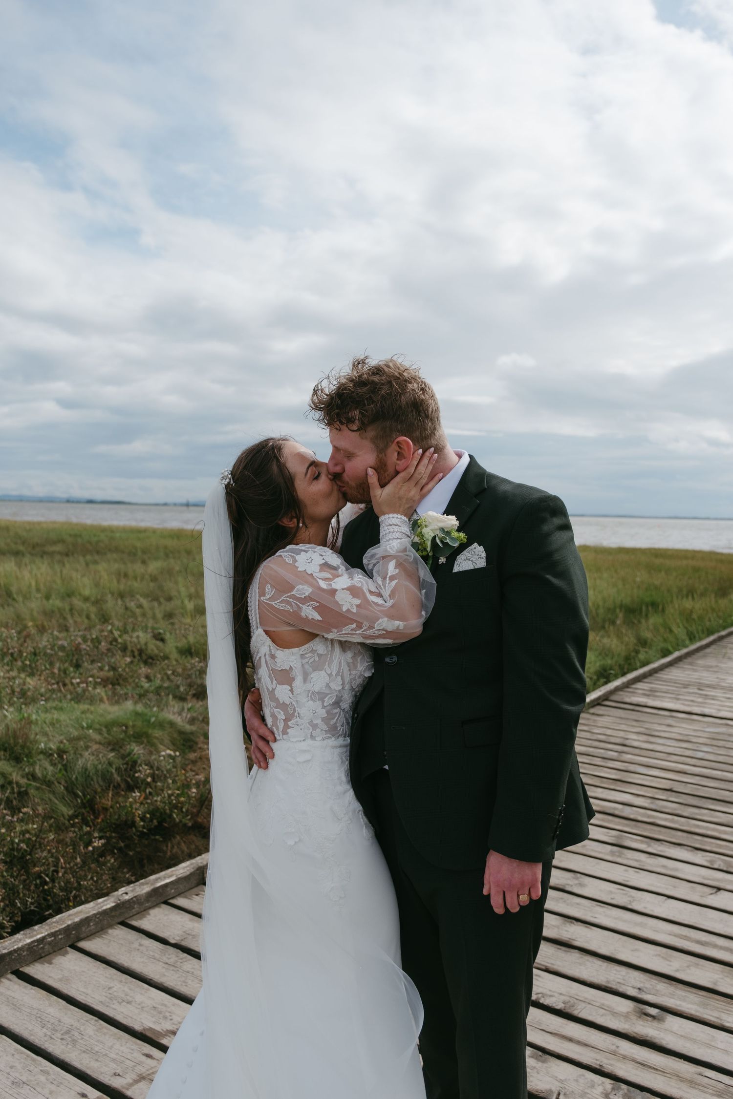 Bride and groom sharing a romantic kiss on a wooden boardwalk surrounded by green marshlands under a cloudy sky.