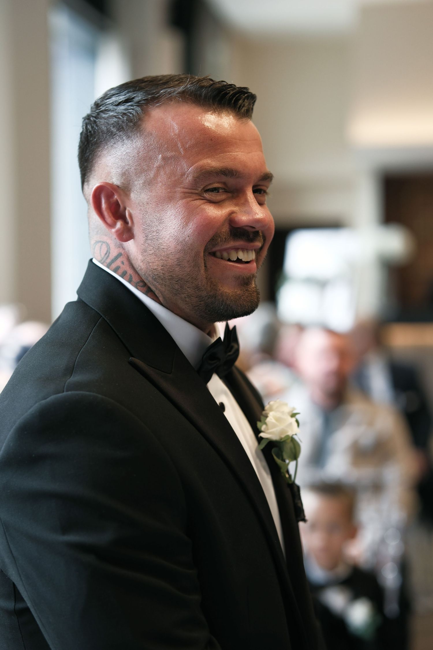 Smiling groom in black tuxedo with white boutonniere awaiting his bride at the wedding altar.
