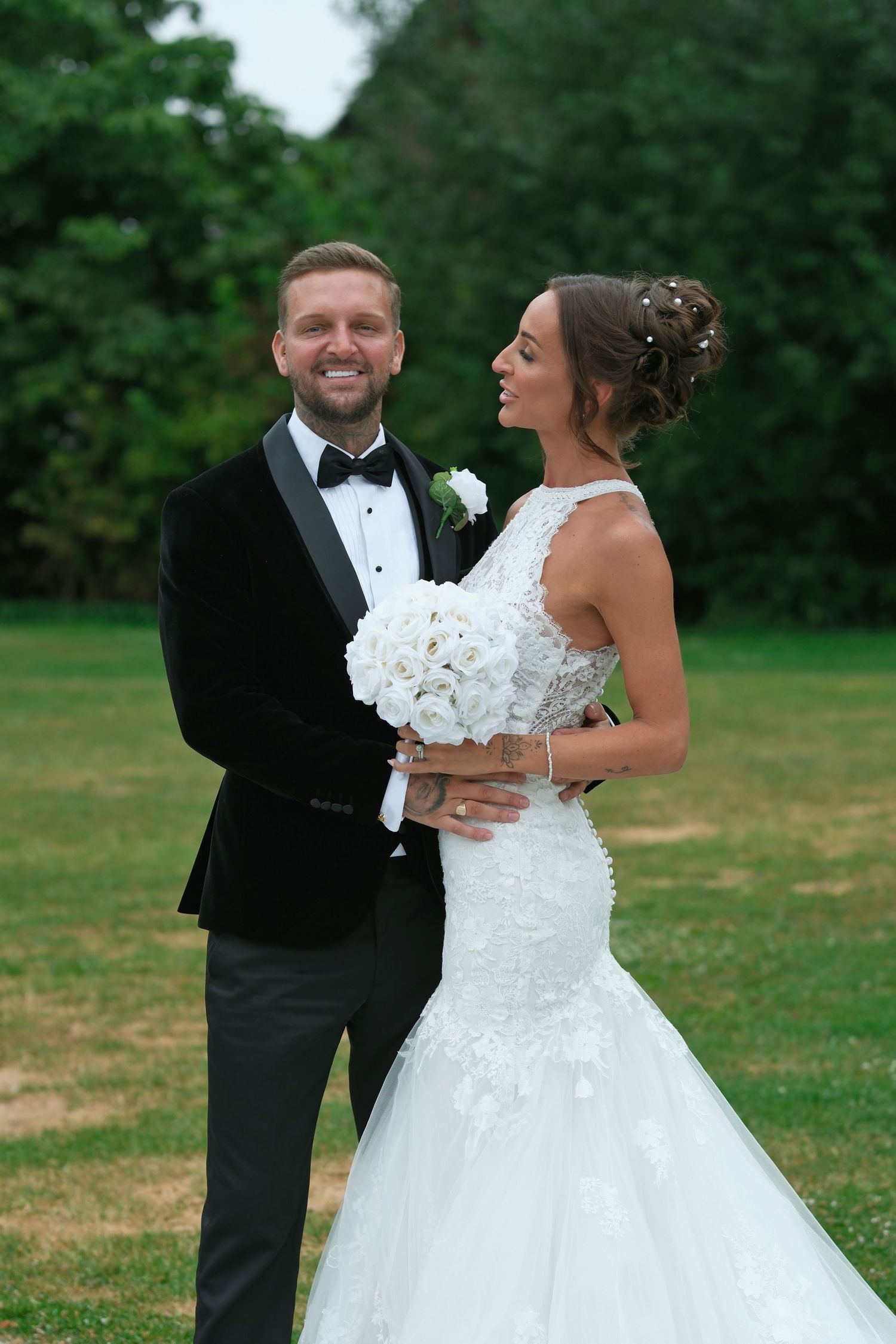A smiling groom in a black tuxedo holds his bride in a white lace mermaid gown with a white bouquet outdoors.