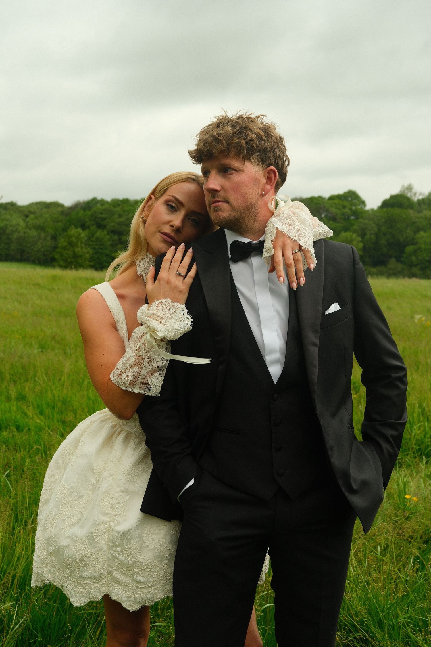 Stylish couple in wedding attire posing intimately in a lush green countryside field.