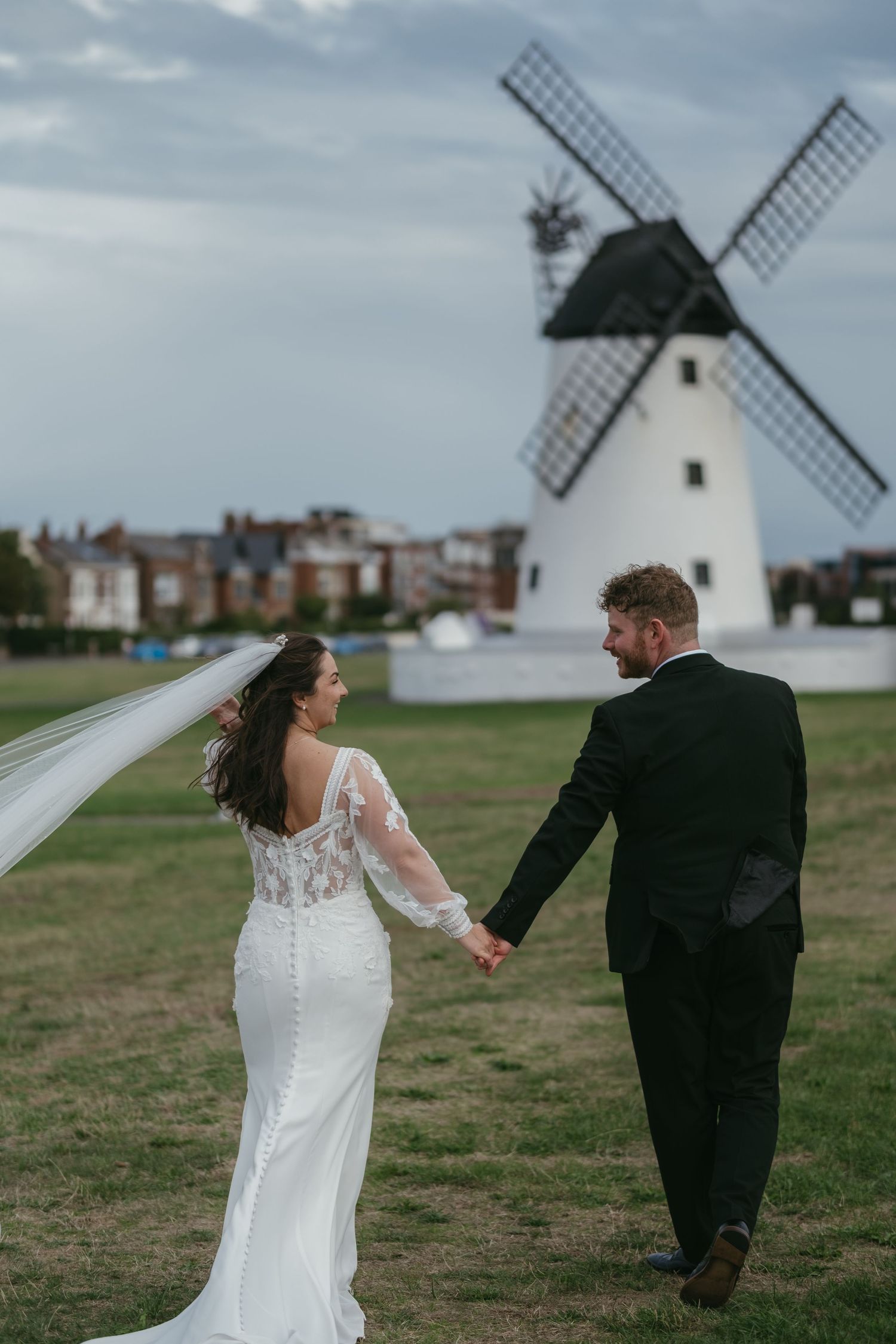 Bride and groom holding hands walking near white windmill on cloudy day, veil blowing in wind.