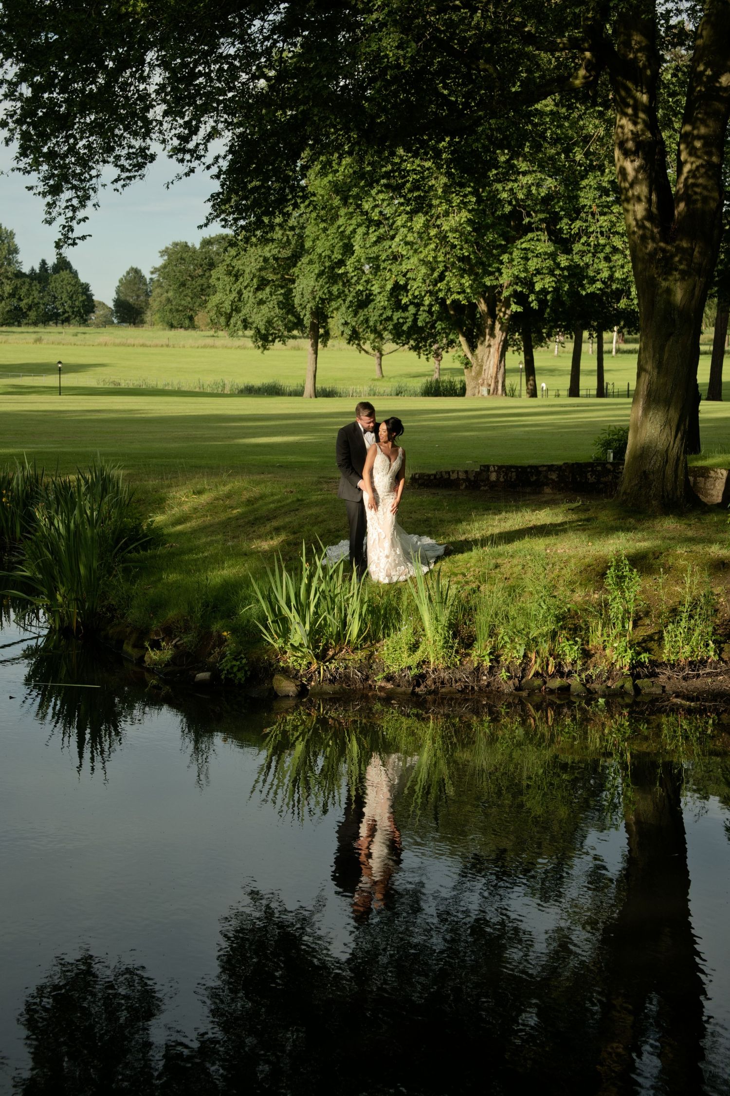 Bride and groom share romantic moment by tranquil pond surrounded by lush green trees, their reflection visible in water.
