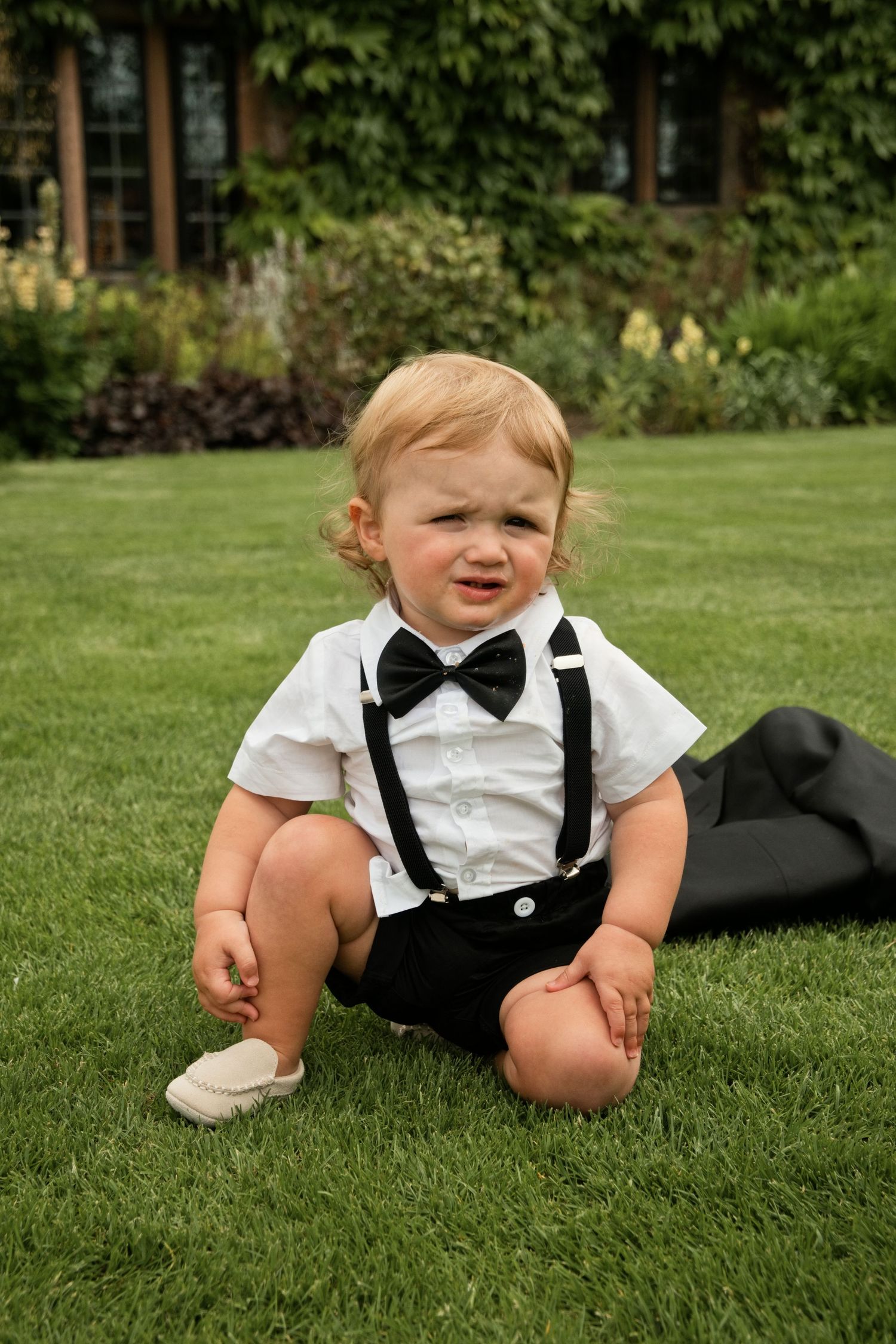 Adorable baby boy in formal black and white outfit with bow tie sitting on green grass.