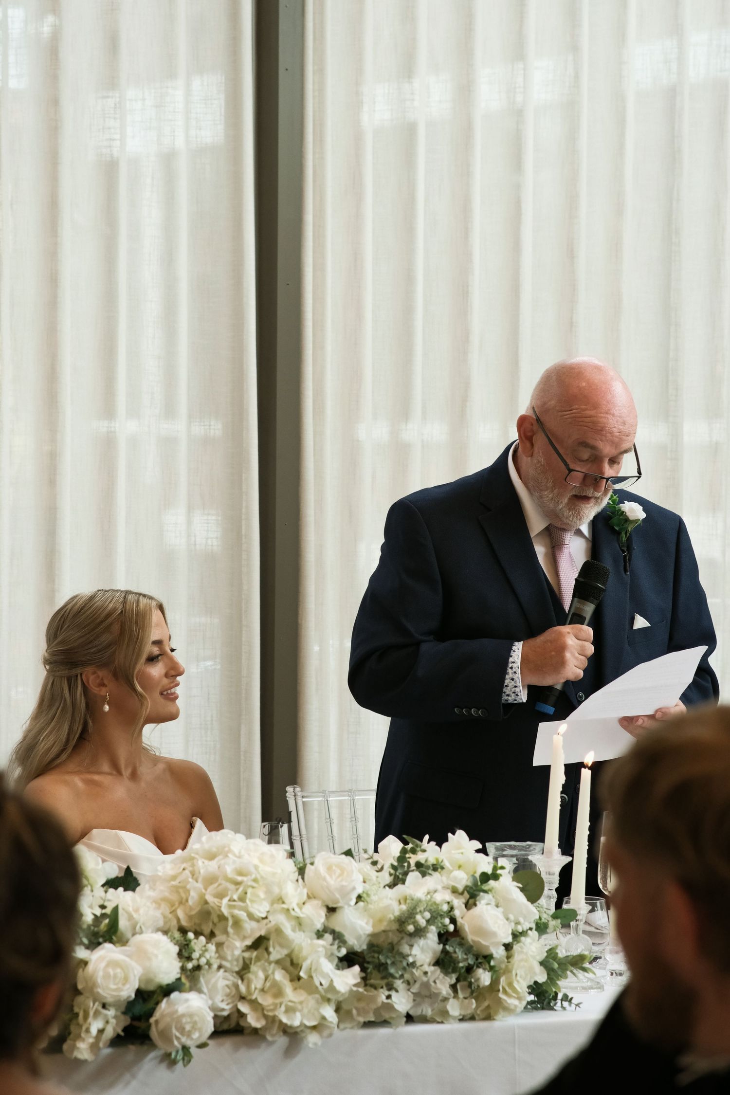 Father of the bride giving speech at elegant wedding reception with white roses.