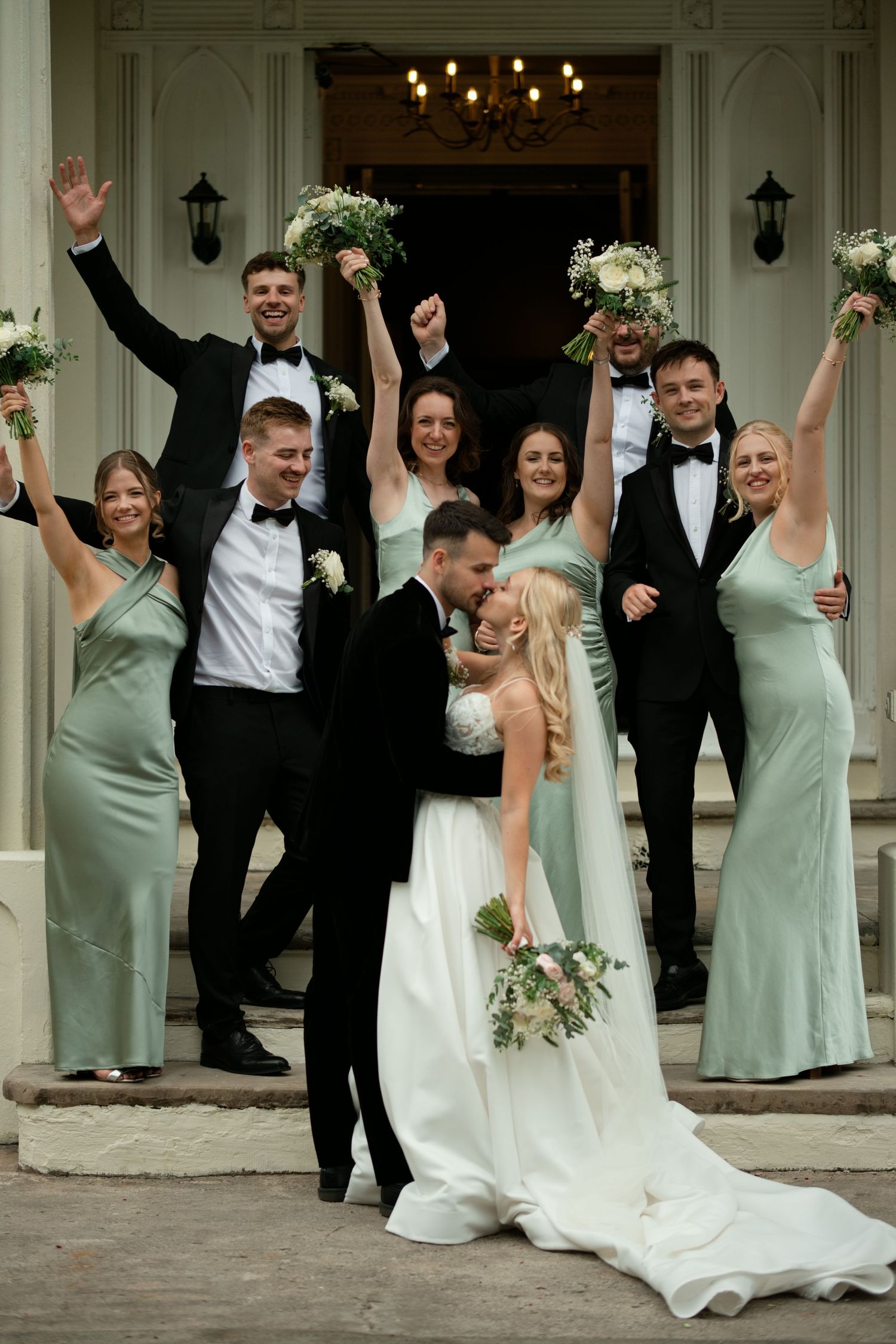 Bride and groom kissing on steps surrounded by cheering wedding party in sage green dresses and black tuxedos.