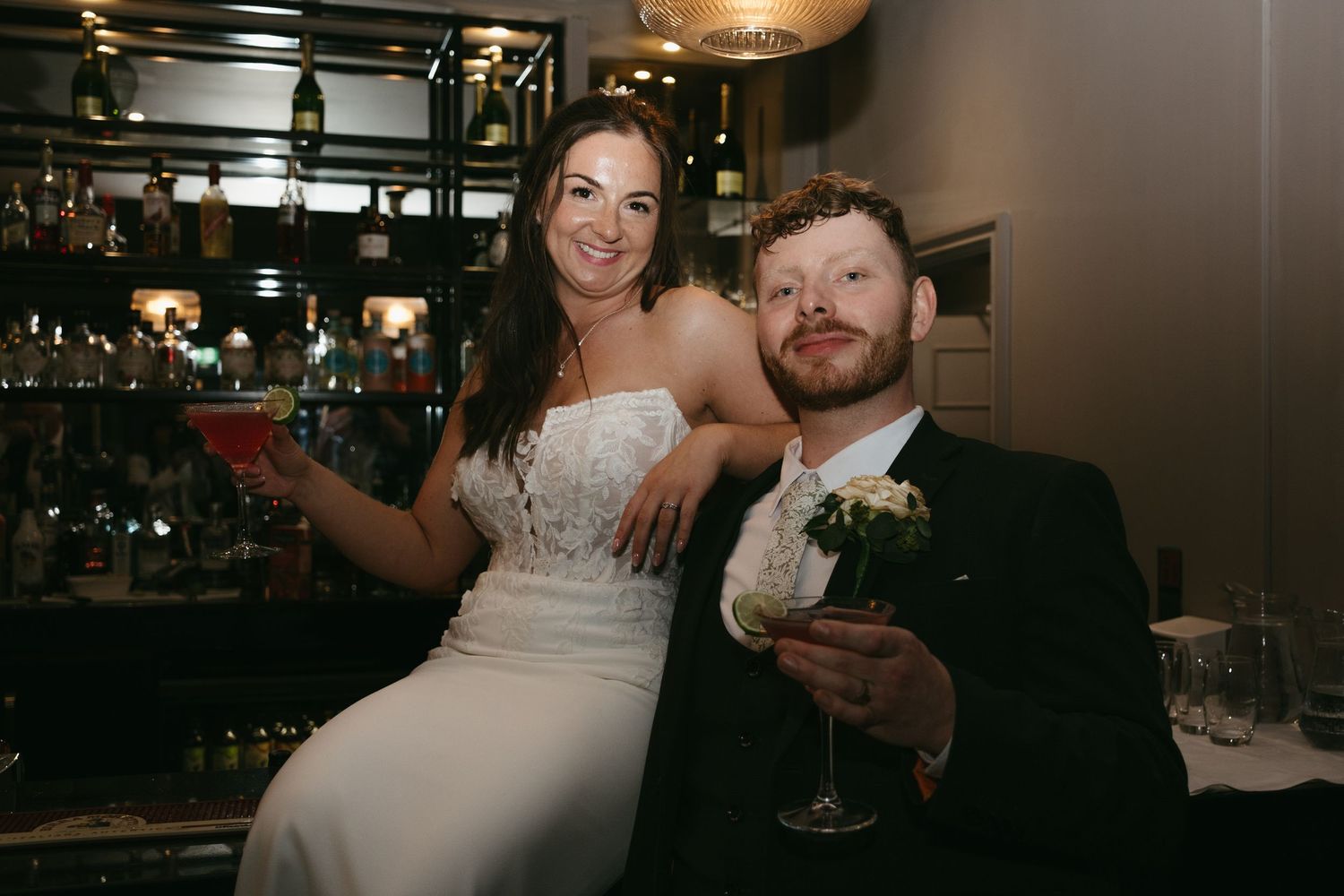 Bride and groom smiling at bar holding cocktails on wedding day.