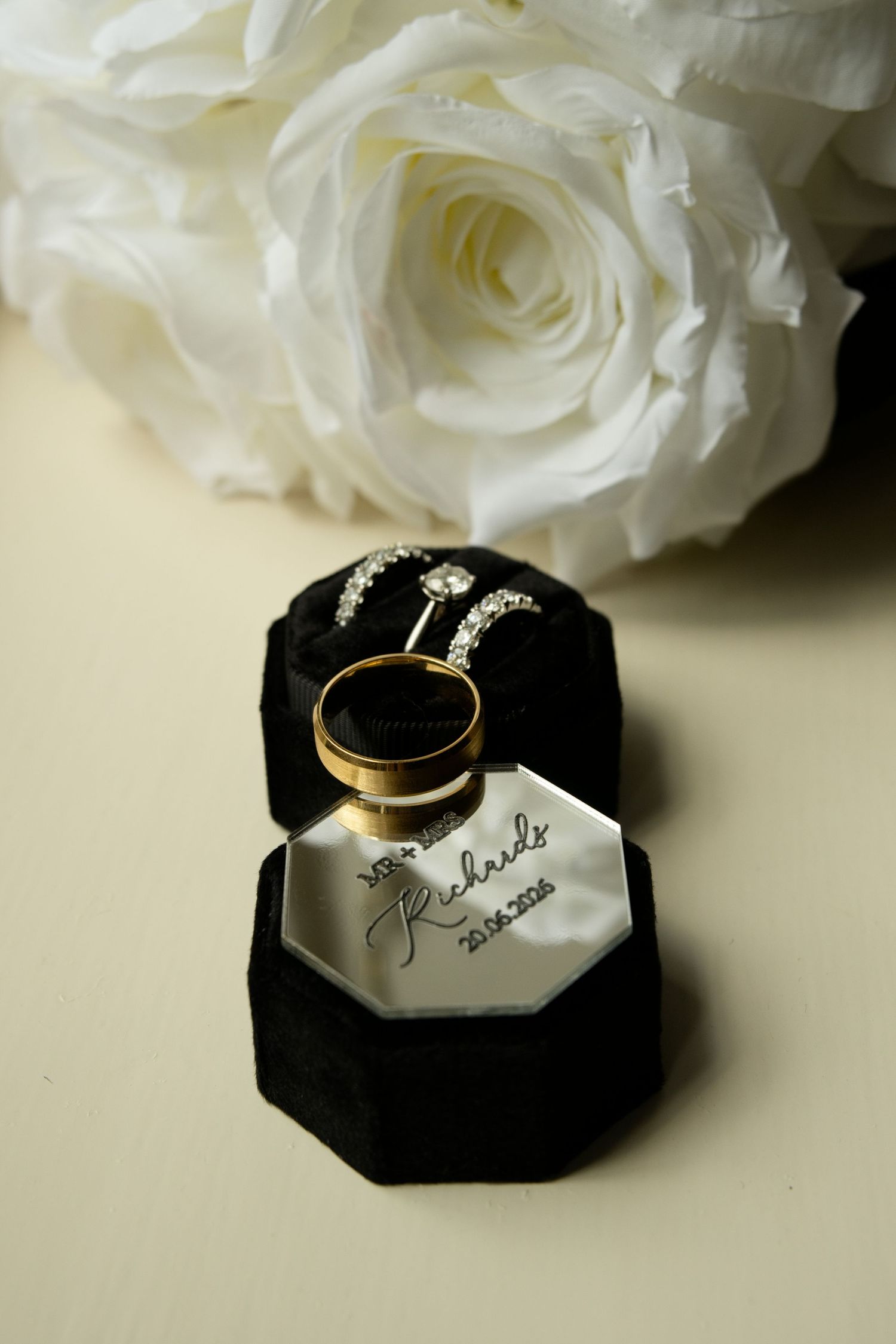 Gold wedding band and diamond engagement ring displayed in black velvet box beside white roses with personalized inscription.