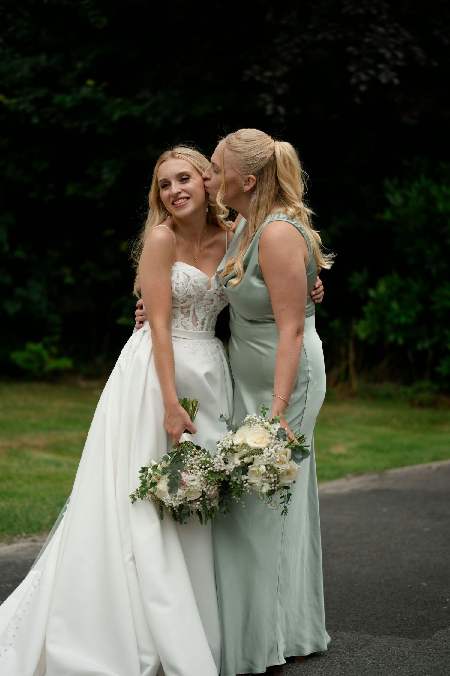 Blonde bride in white lace gown and bridesmaid in sage green dress sharing a tender moment holding bouquets.