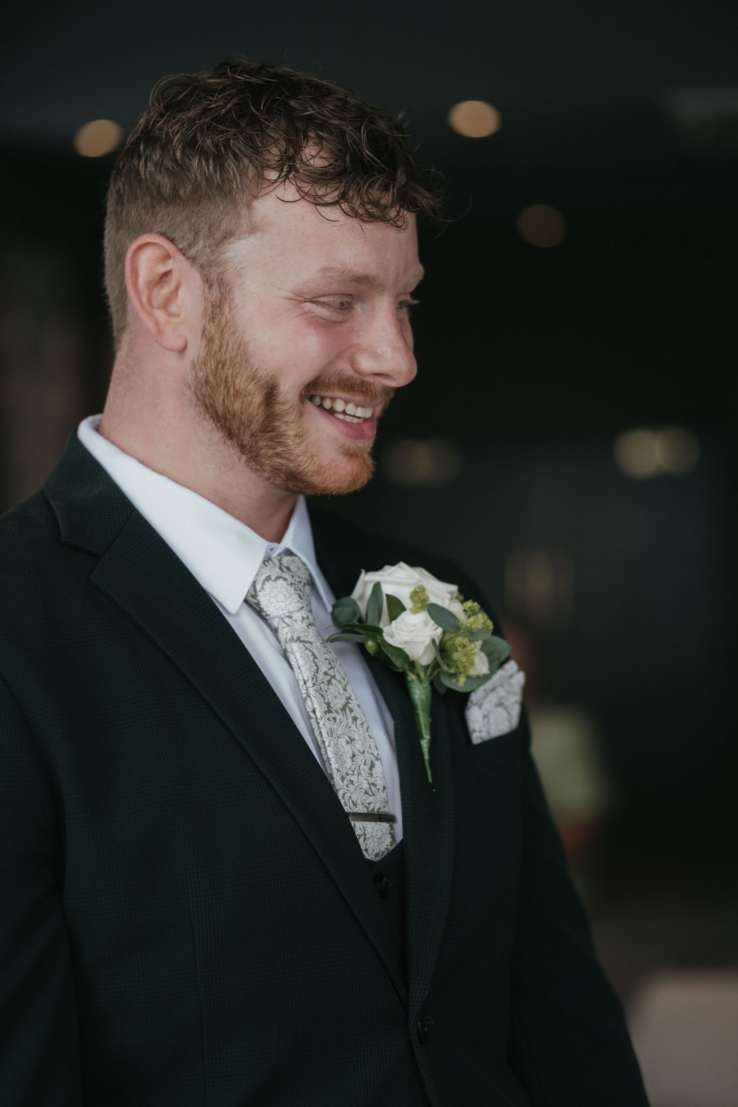 Smiling groom in dark suit with white rose boutonniere and patterned tie on his wedding day.