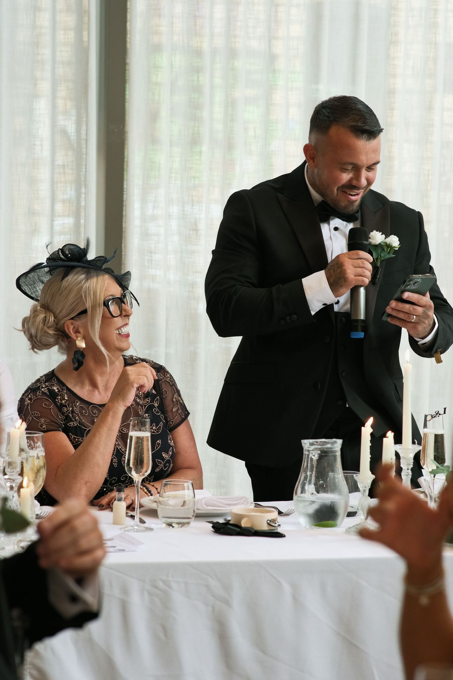Groomsman delivering wedding speech from phone beside smiling woman in fascinator.