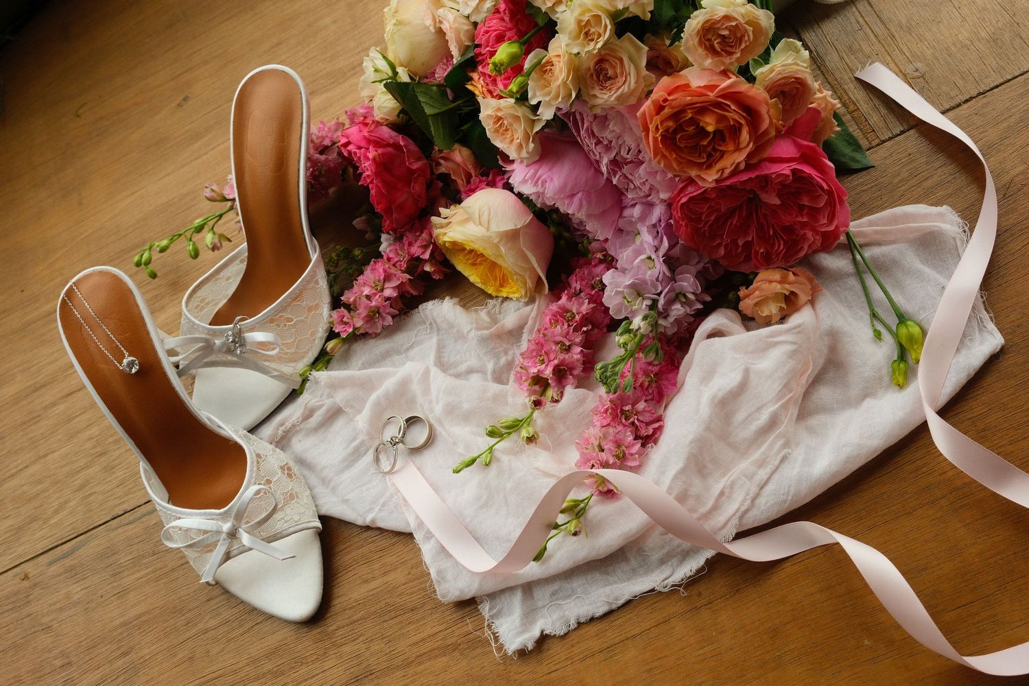 Wedding flat lay featuring white heeled bridal shoes, colorful rose bouquet, and rings on lace fabric.