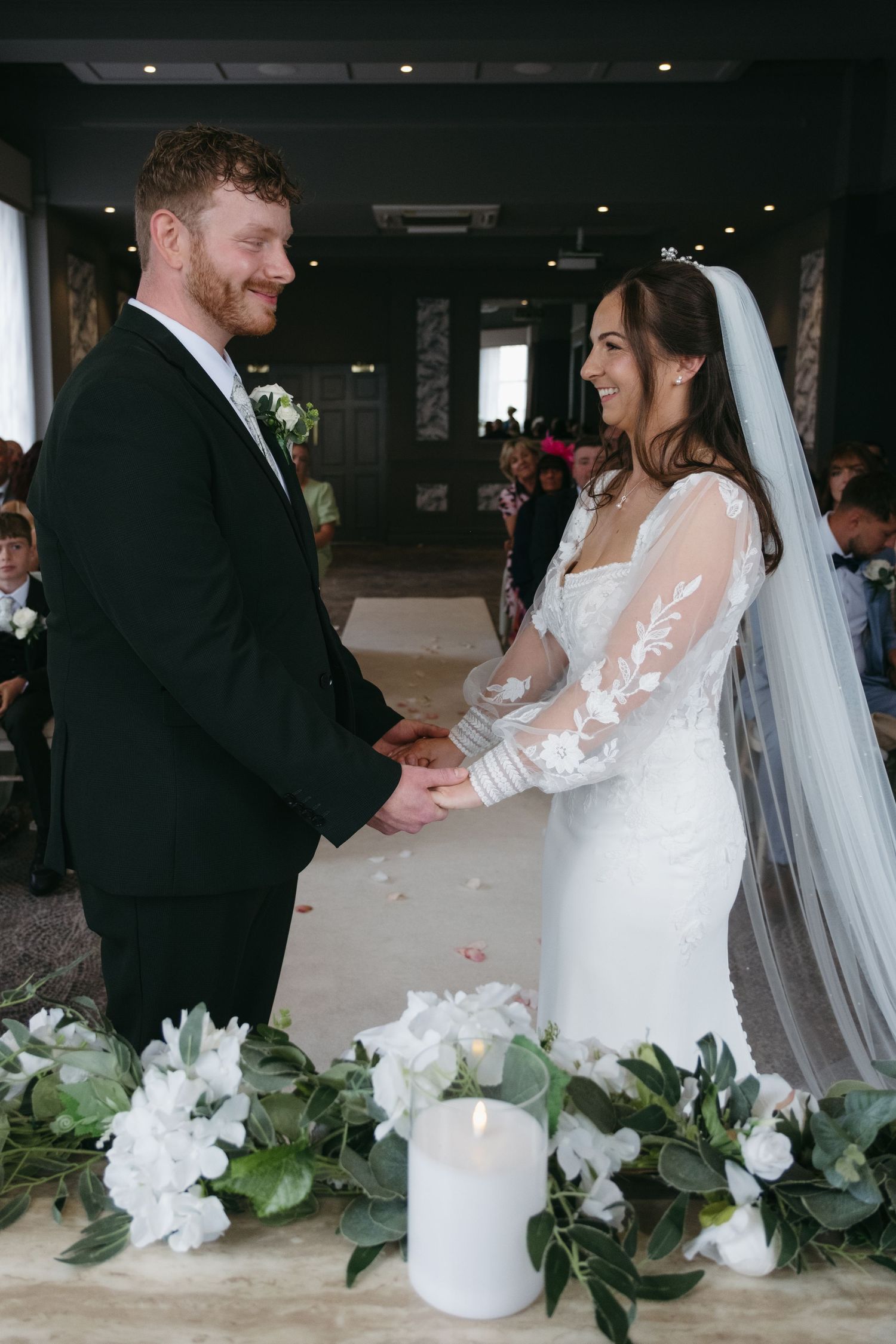 A bride and groom hold hands and smile during their wedding ceremony, with white floral decorations and a candle in the foreground.