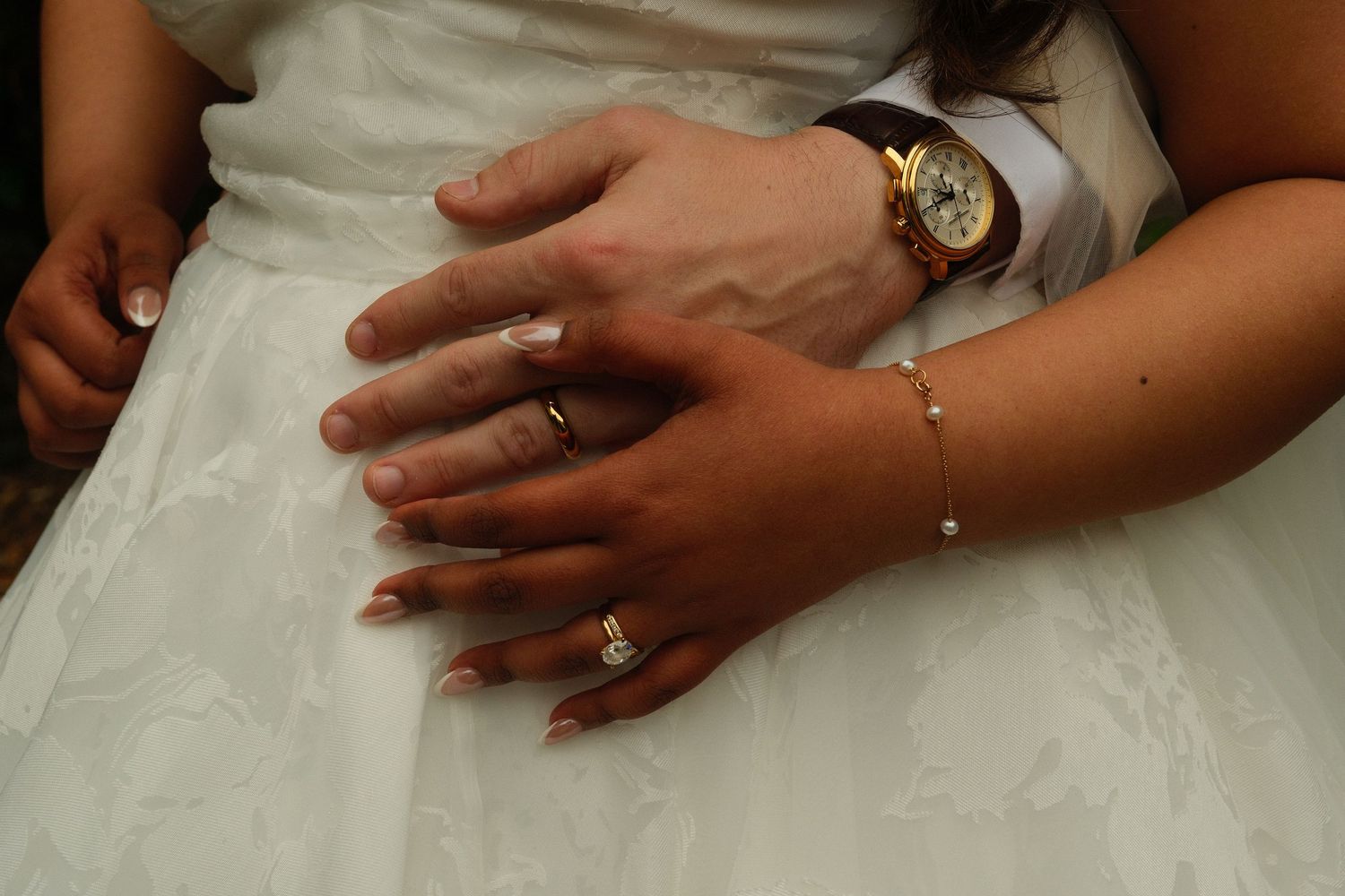 Close-up of newlyweds hands showing gold wedding rings, bracelet, and vintage watch against white bridal gown.