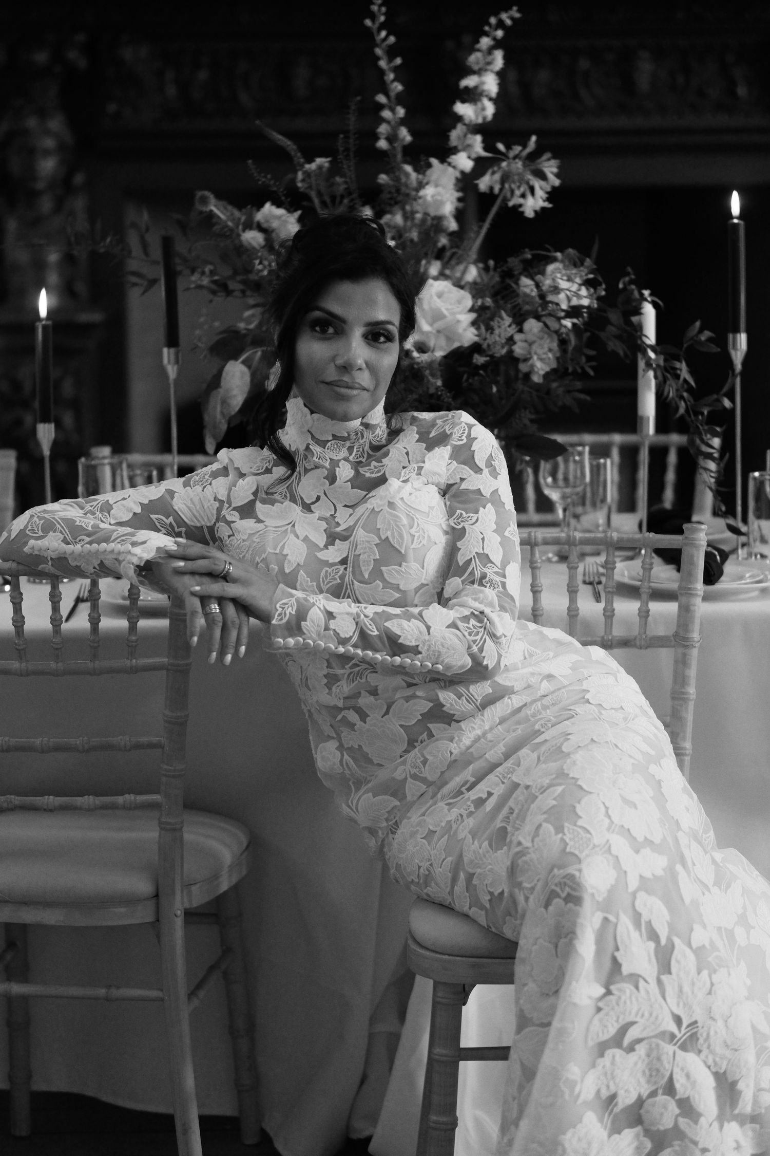 Black and white photo of a bride in lace gown seated at an elegant candlelit reception table with floral arrangements.