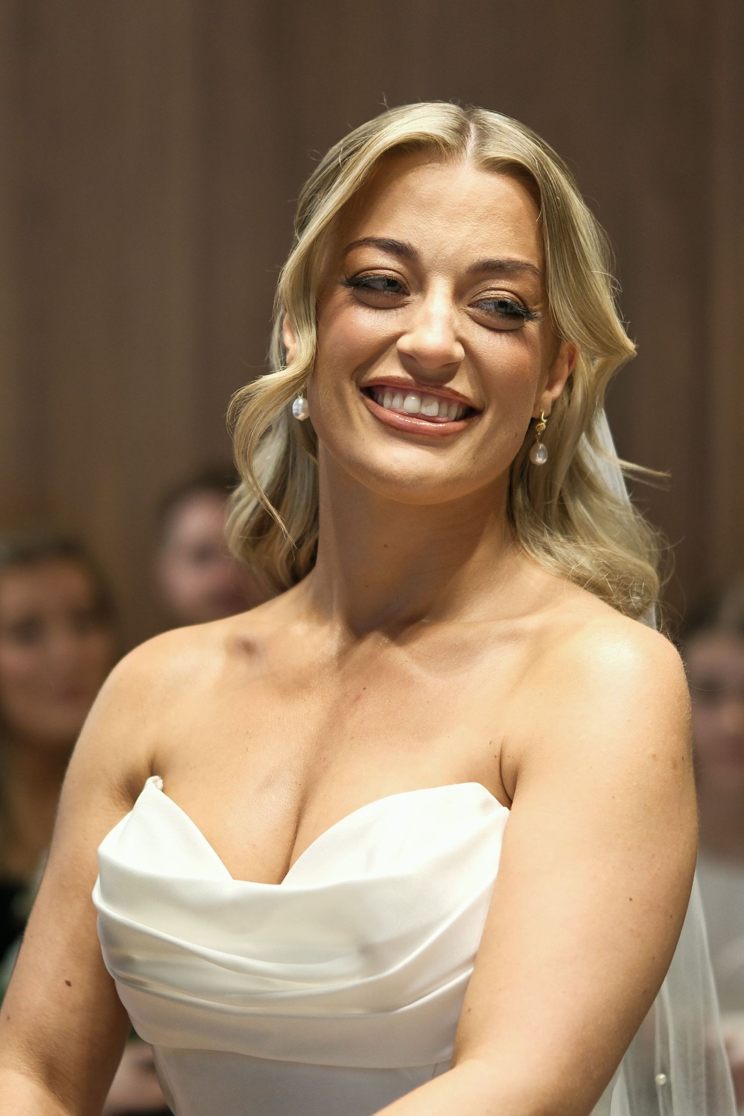 Radiant blonde bride in strapless white gown smiles joyfully during wedding ceremony.