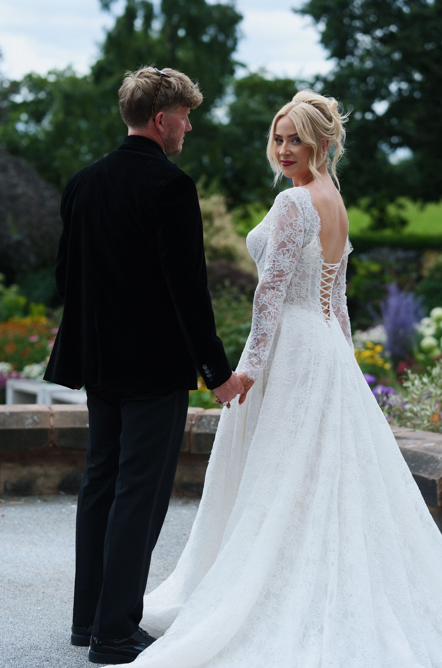 Bride in lace long-sleeve wedding gown with corset back holds hands with groom in black suit outdoors.