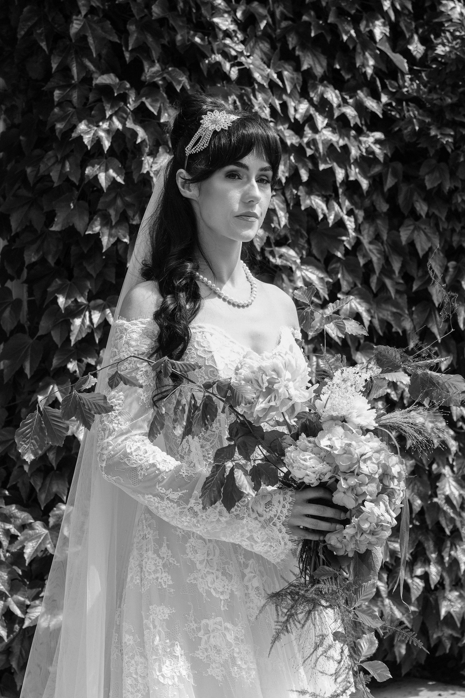 Black and white photo of a bride in lace gown holding a bouquet, wearing a veil and pearl necklace before ivy wall.