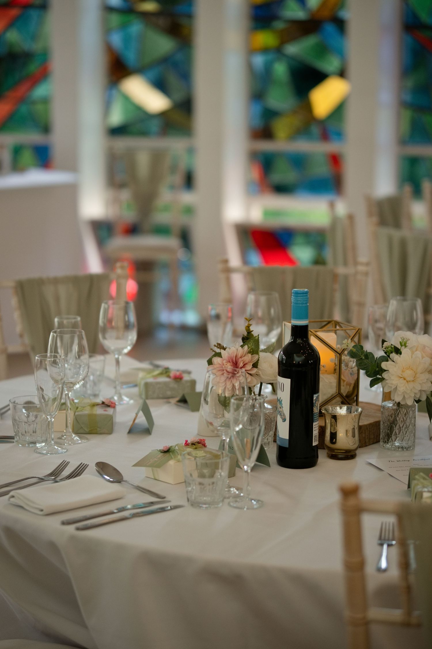 Elegantly set wedding reception table with white linens, crystal glasses, wine bottle and stained glass windows.