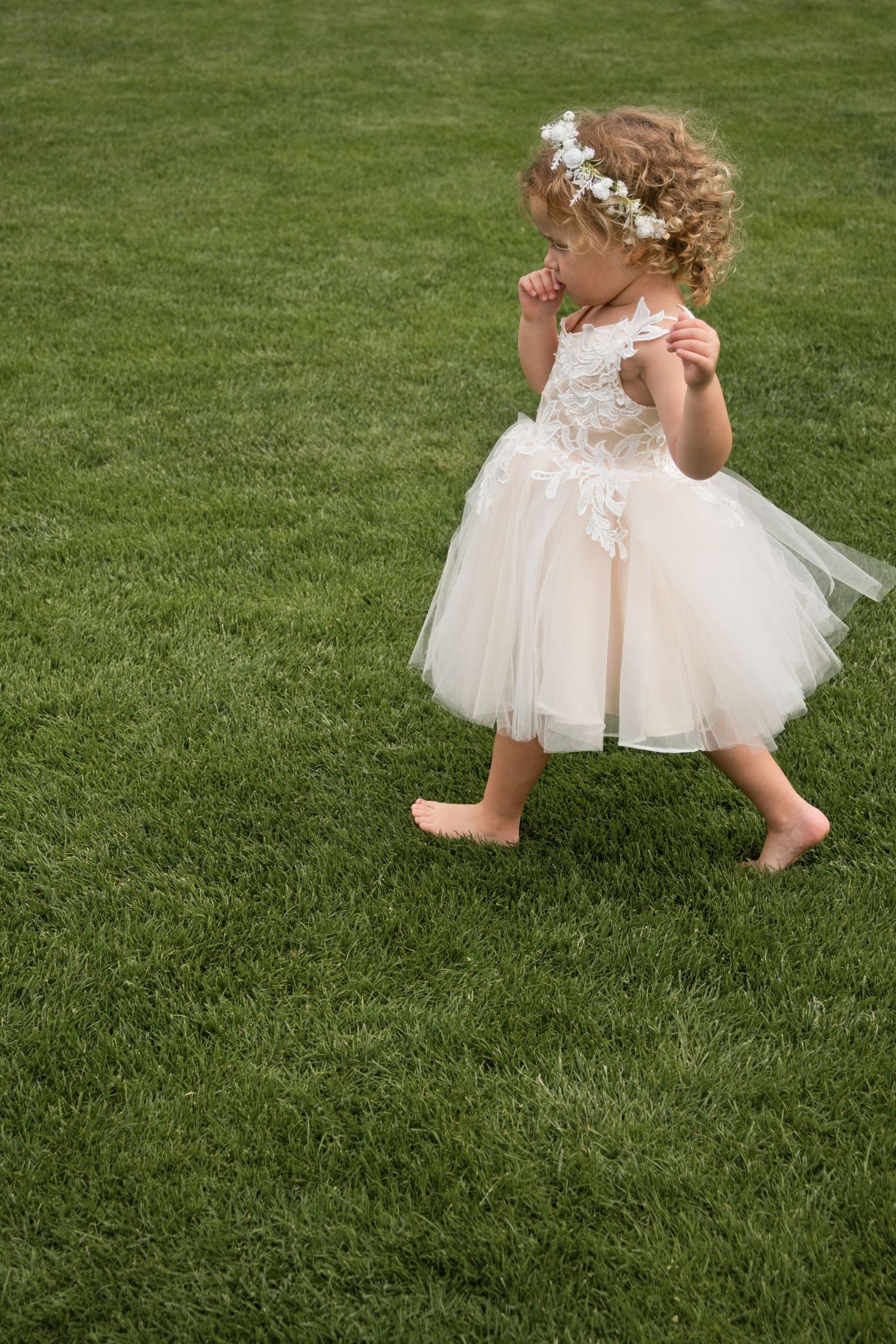 Sweet barefoot flower girl in ivory tulle dress with floral crown walking on lush green lawn.