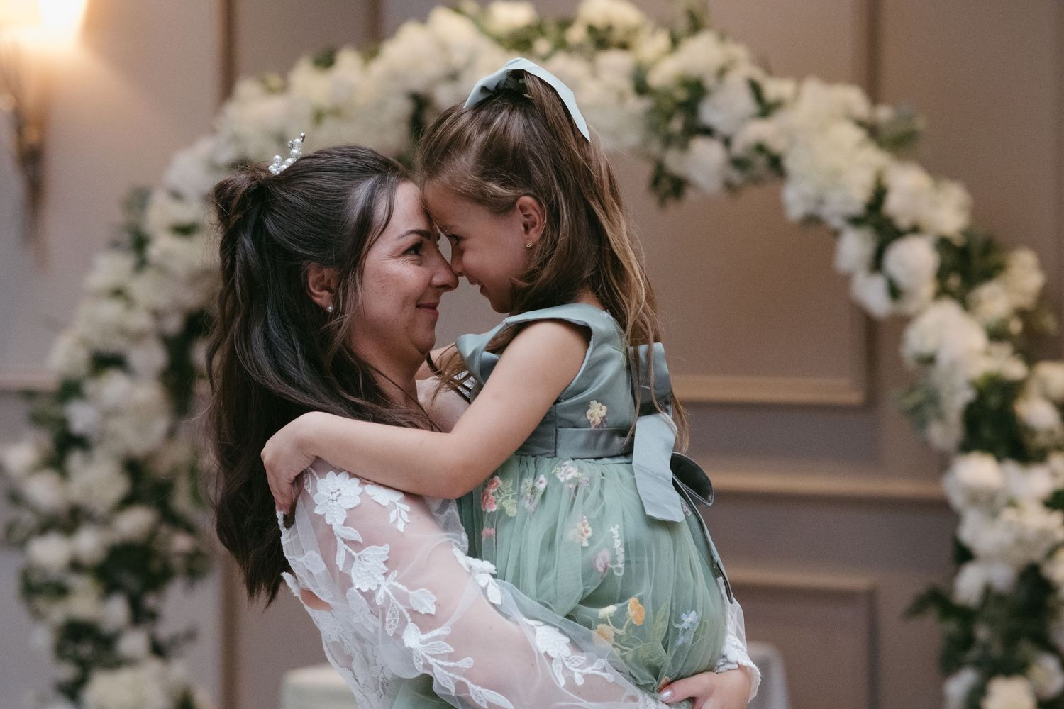 A mother in a white lace gown holds her young daughter in a mint dress, sharing a tender moment near a floral arch.