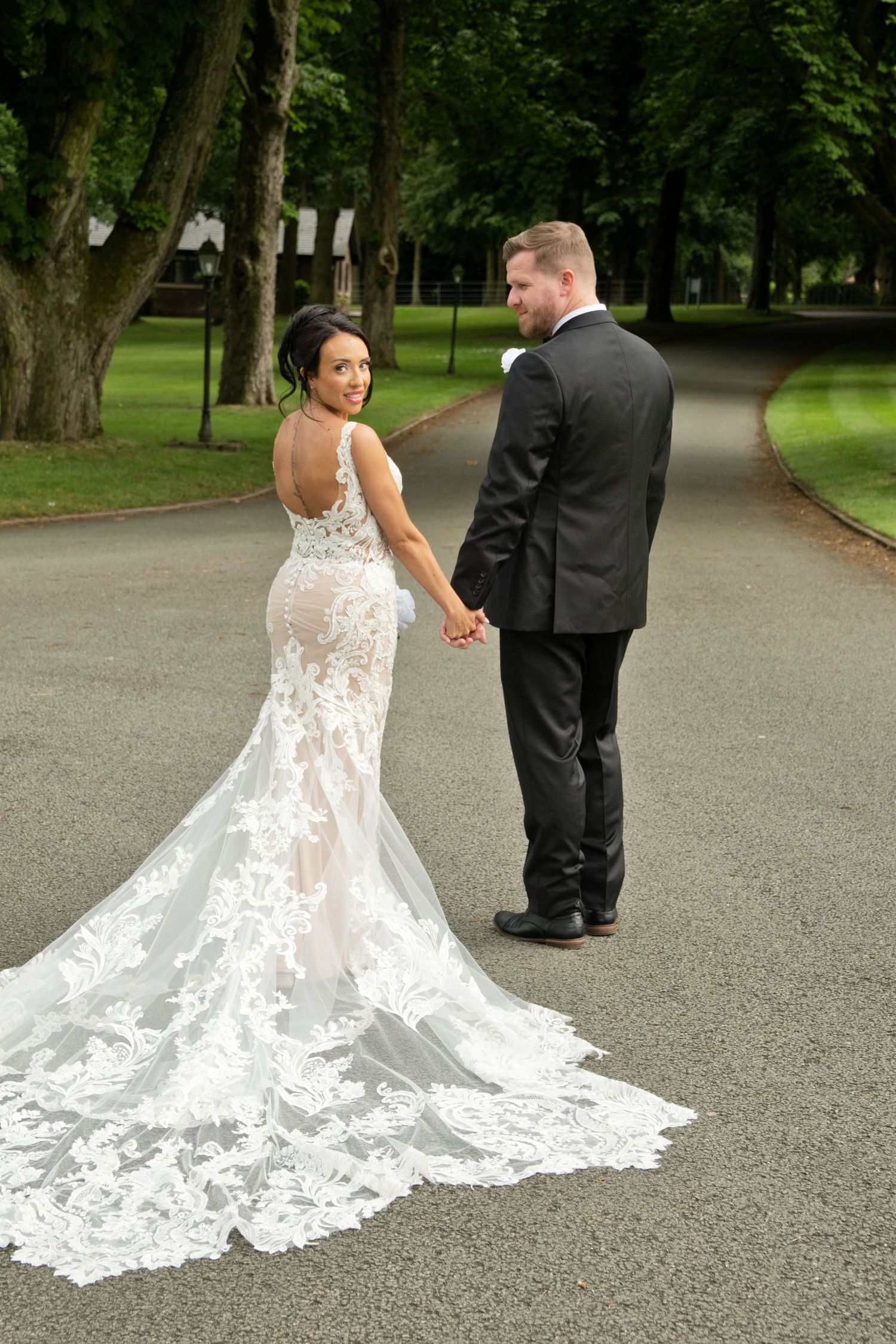 Bride in backless lace gown with long train holds grooms hand on tree-lined driveway, looking back at camera.