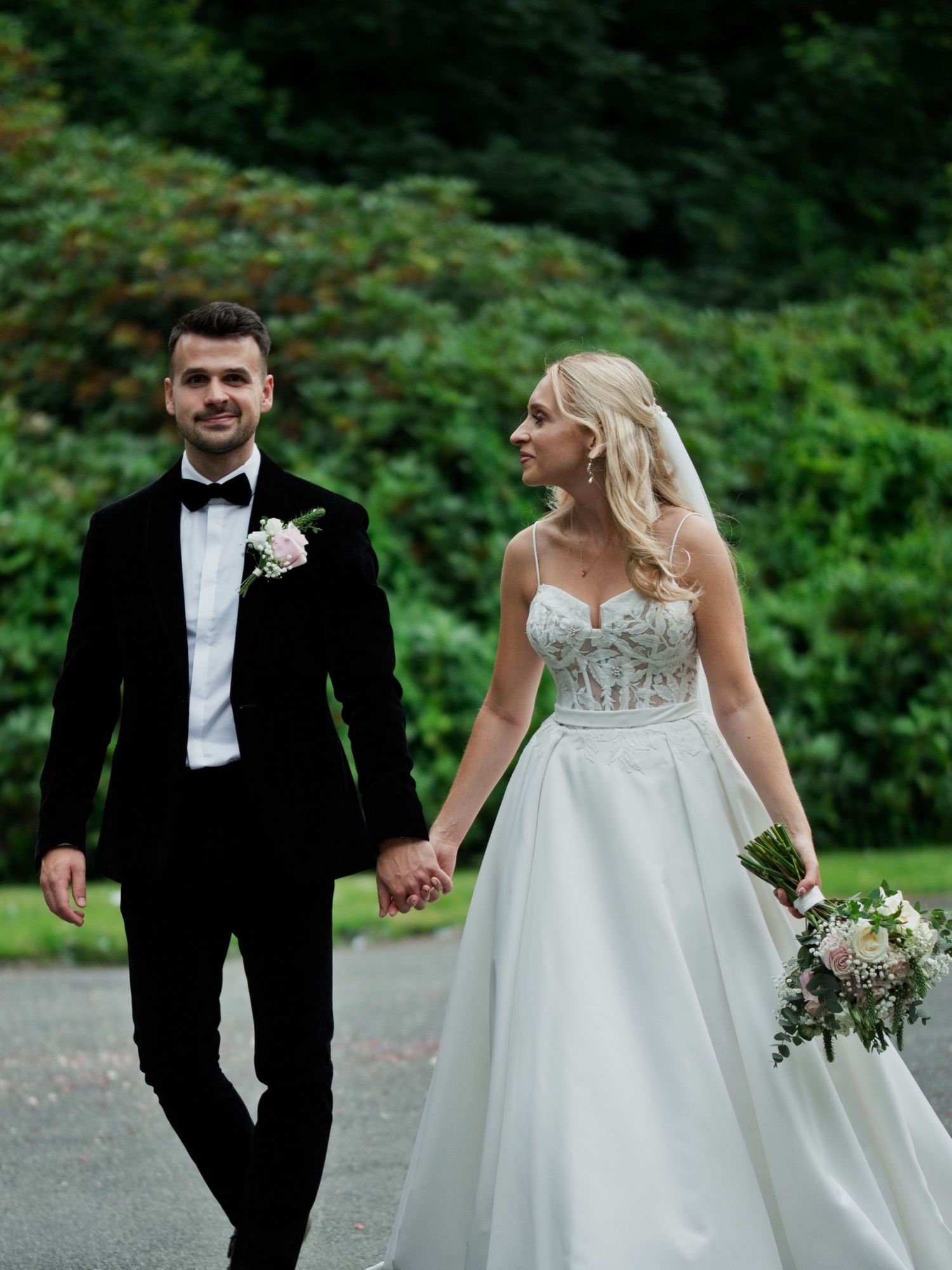 Happy bride and groom walk hand in hand through beautiful green gardens on their wedding day.