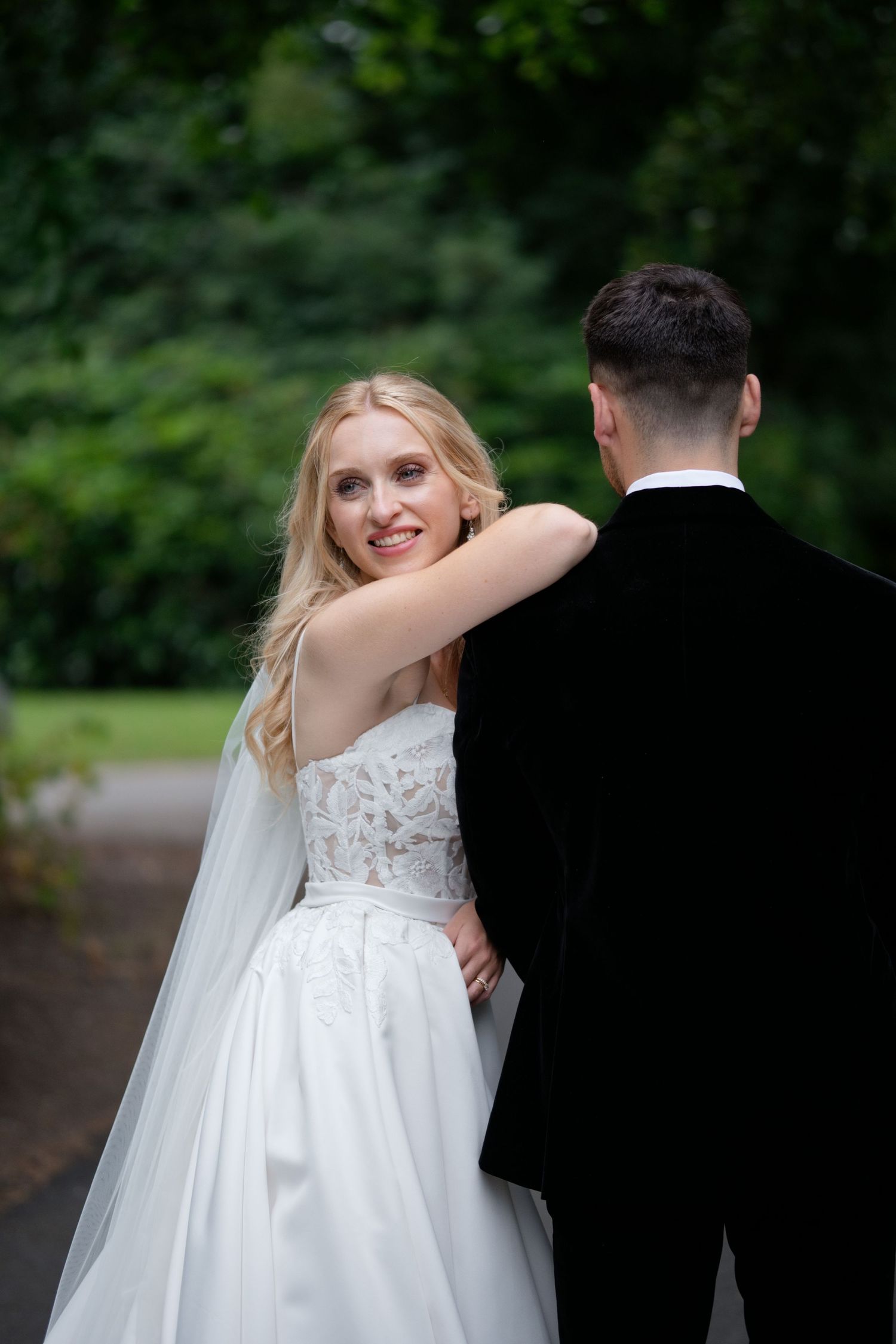 Blonde bride in white lace gown smiling over shoulder while embracing groom in black suit in garden setting.