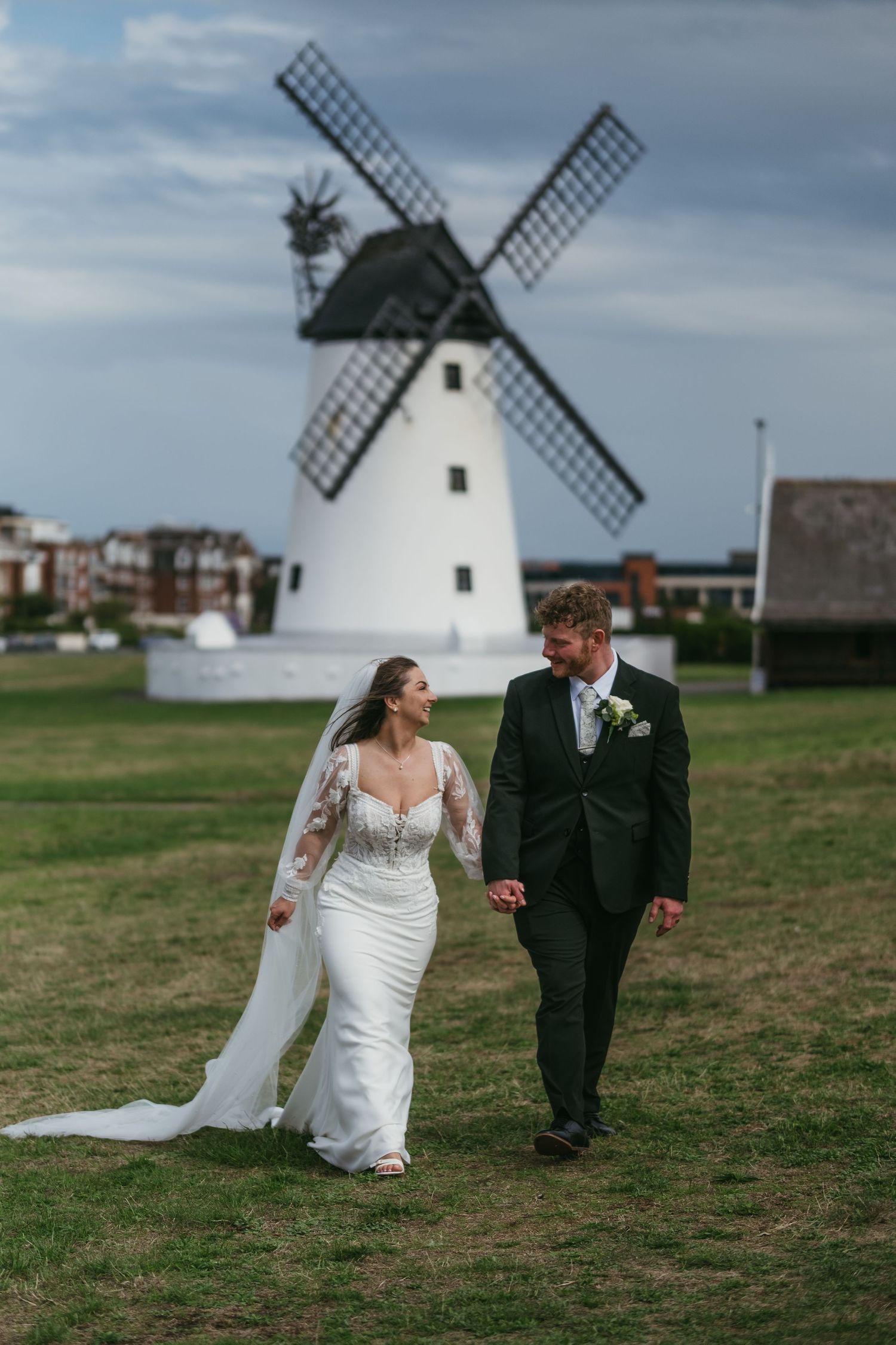 A bride and groom walk hand-in-hand on green grass with a white windmill backdrop on their wedding day.