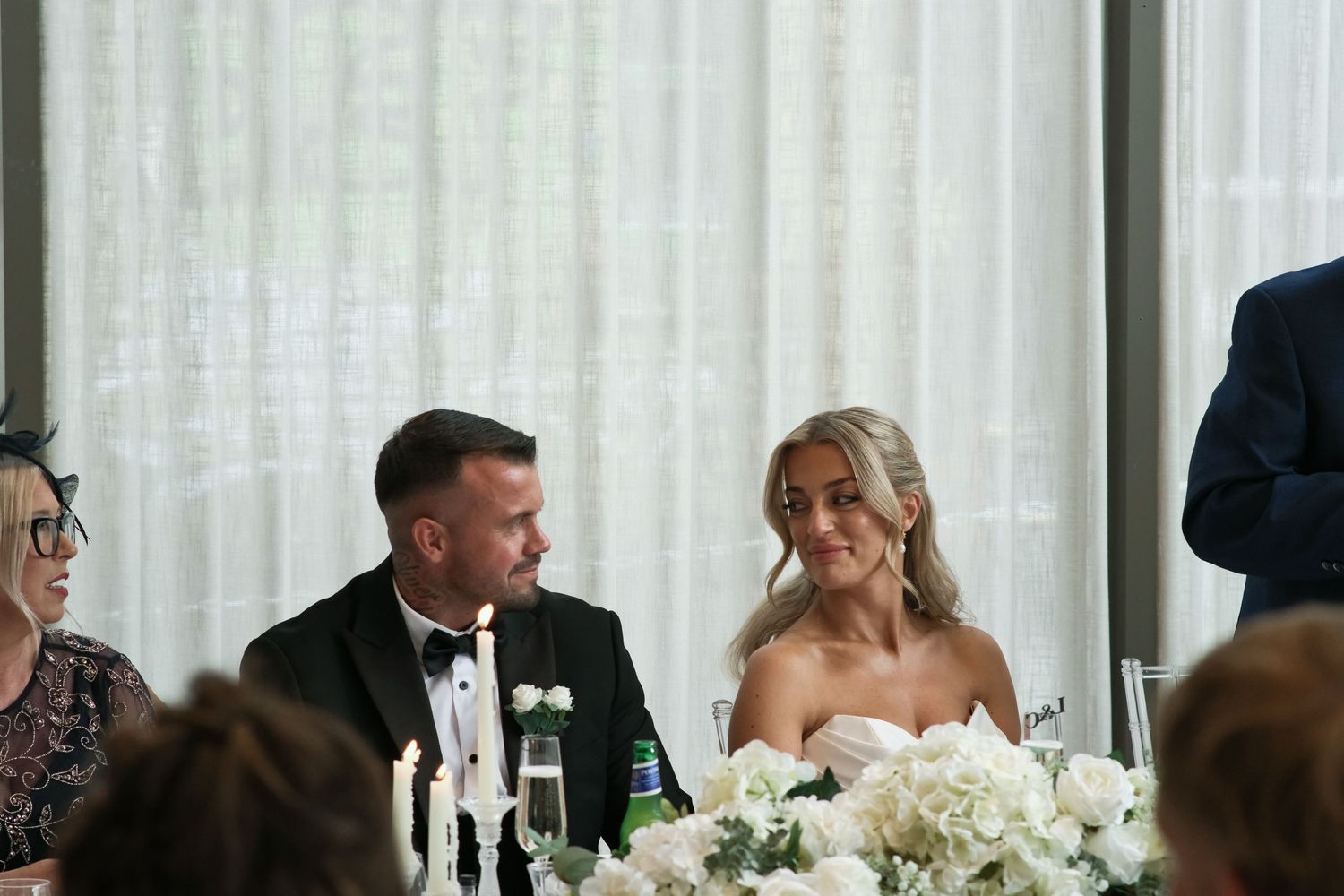 Bride and groom smiling at wedding reception table with white floral centerpiece.