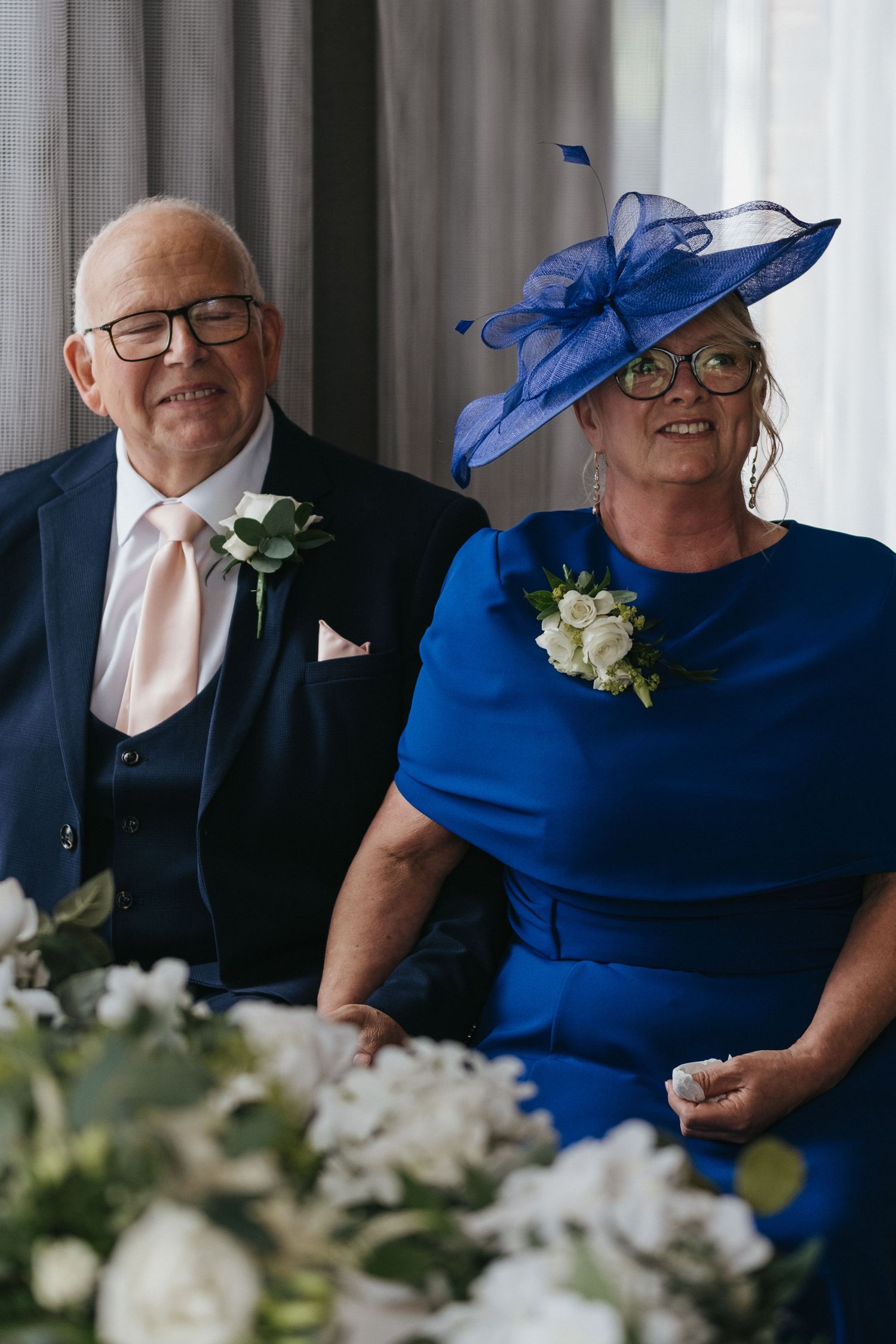 Elderly couple in formal wedding attire, man in navy suit, woman in royal blue dress and matching fascinator hat.