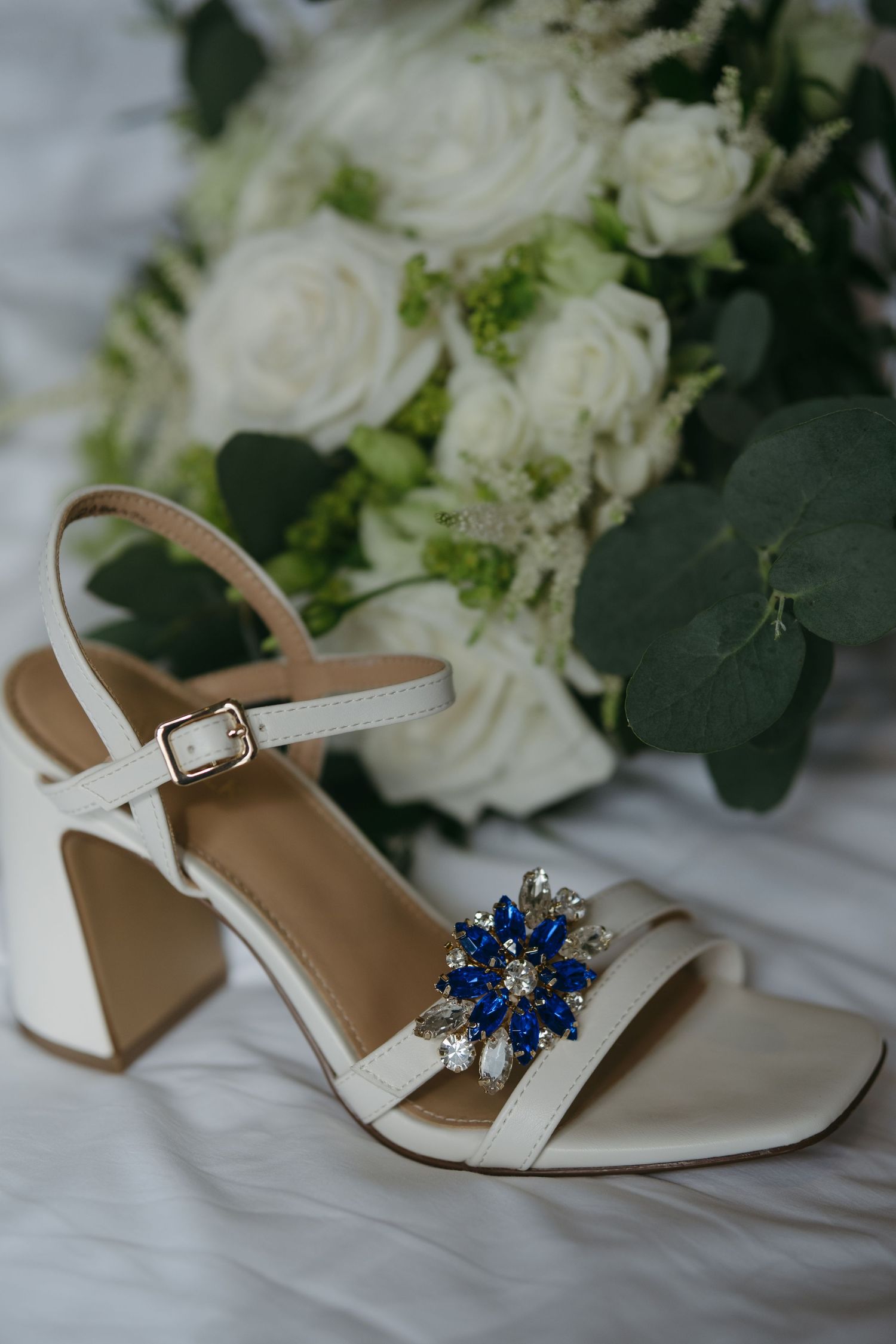 White bridal heels with blue crystal brooch beside white rose wedding bouquet on white fabric.