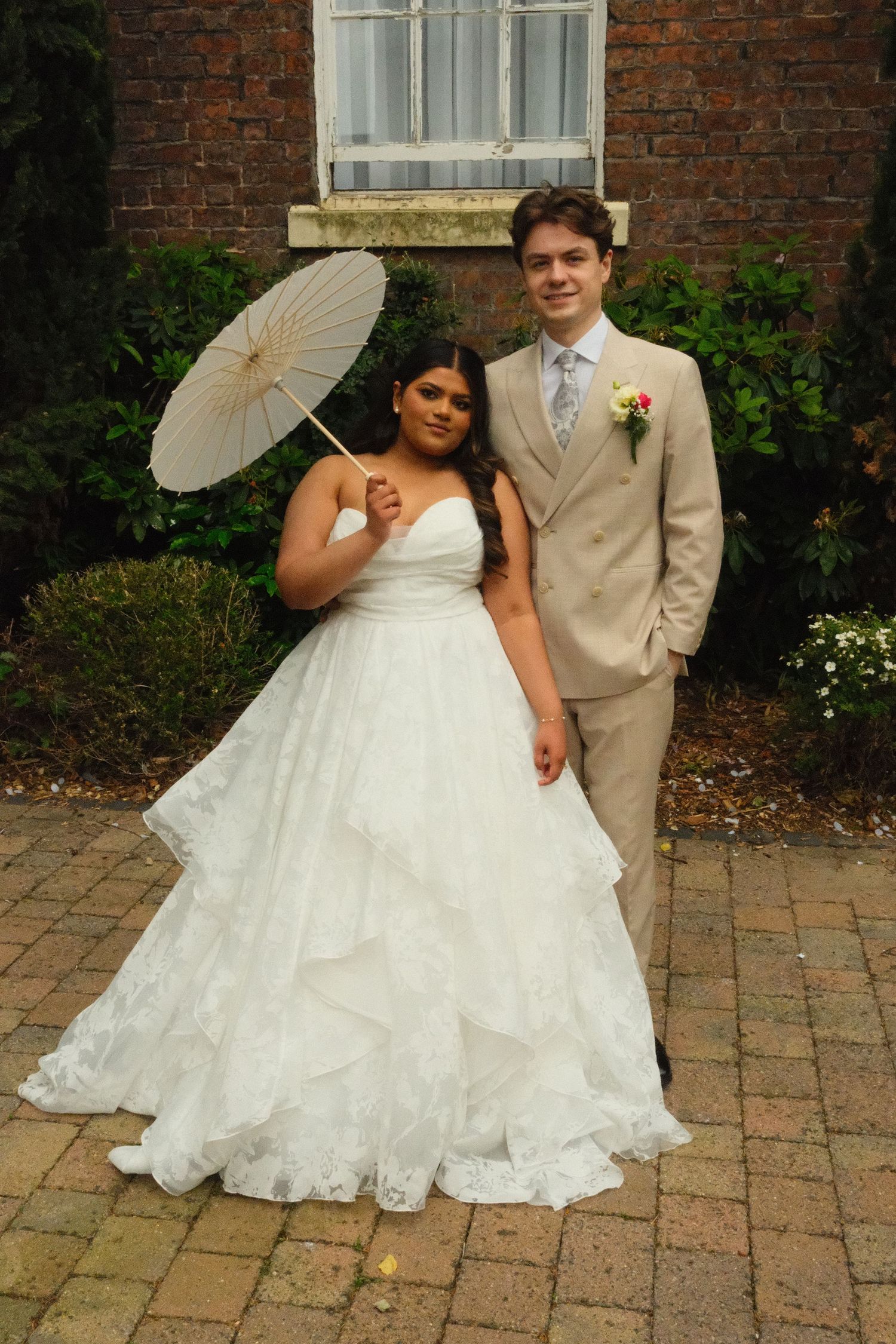Bride in layered white gown holding parasol poses with groom in tan suit against brick wall backdrop.