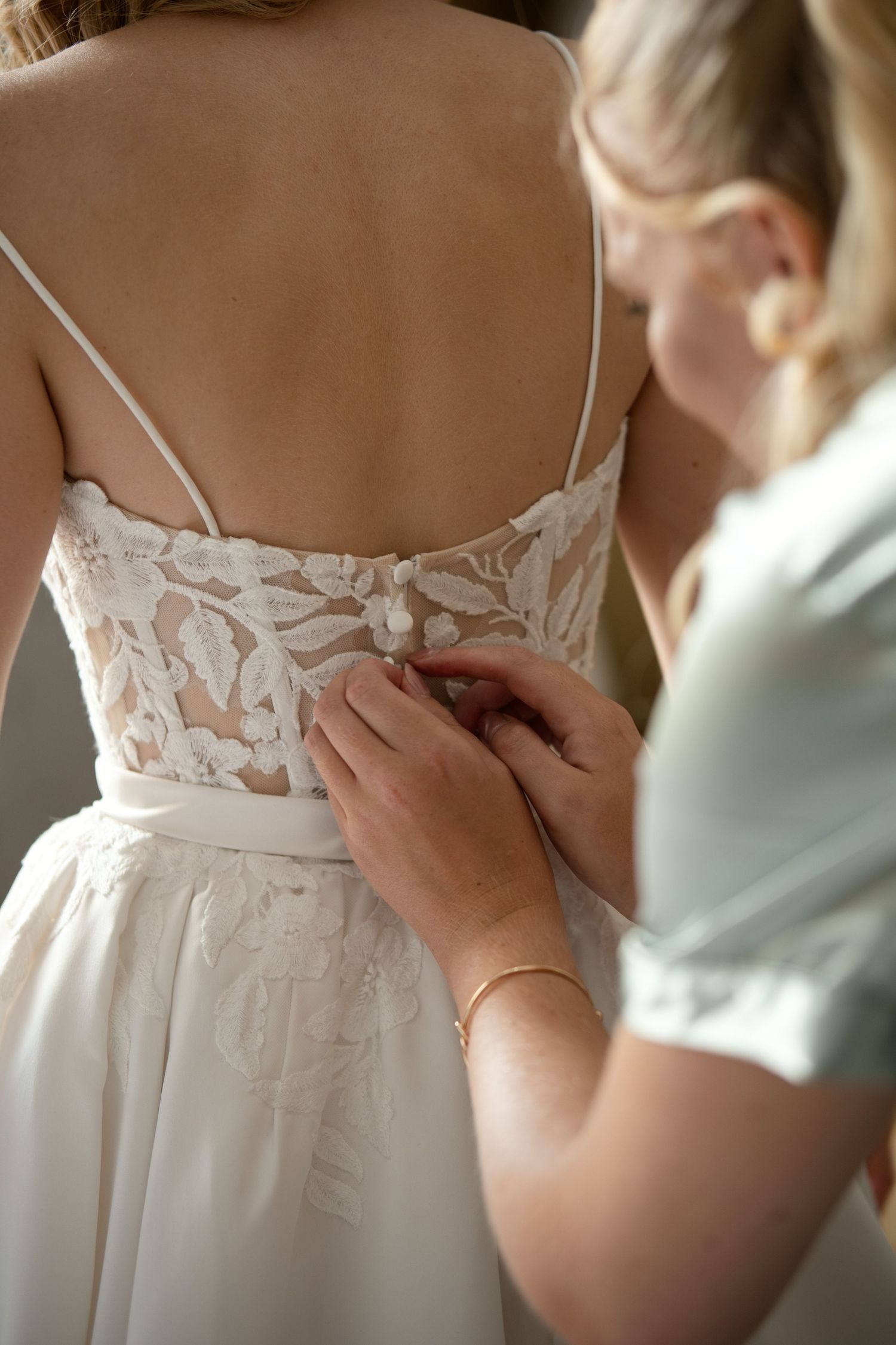 Bridesmaid fastening buttons on back of bride's lace wedding dress during bridal preparations.