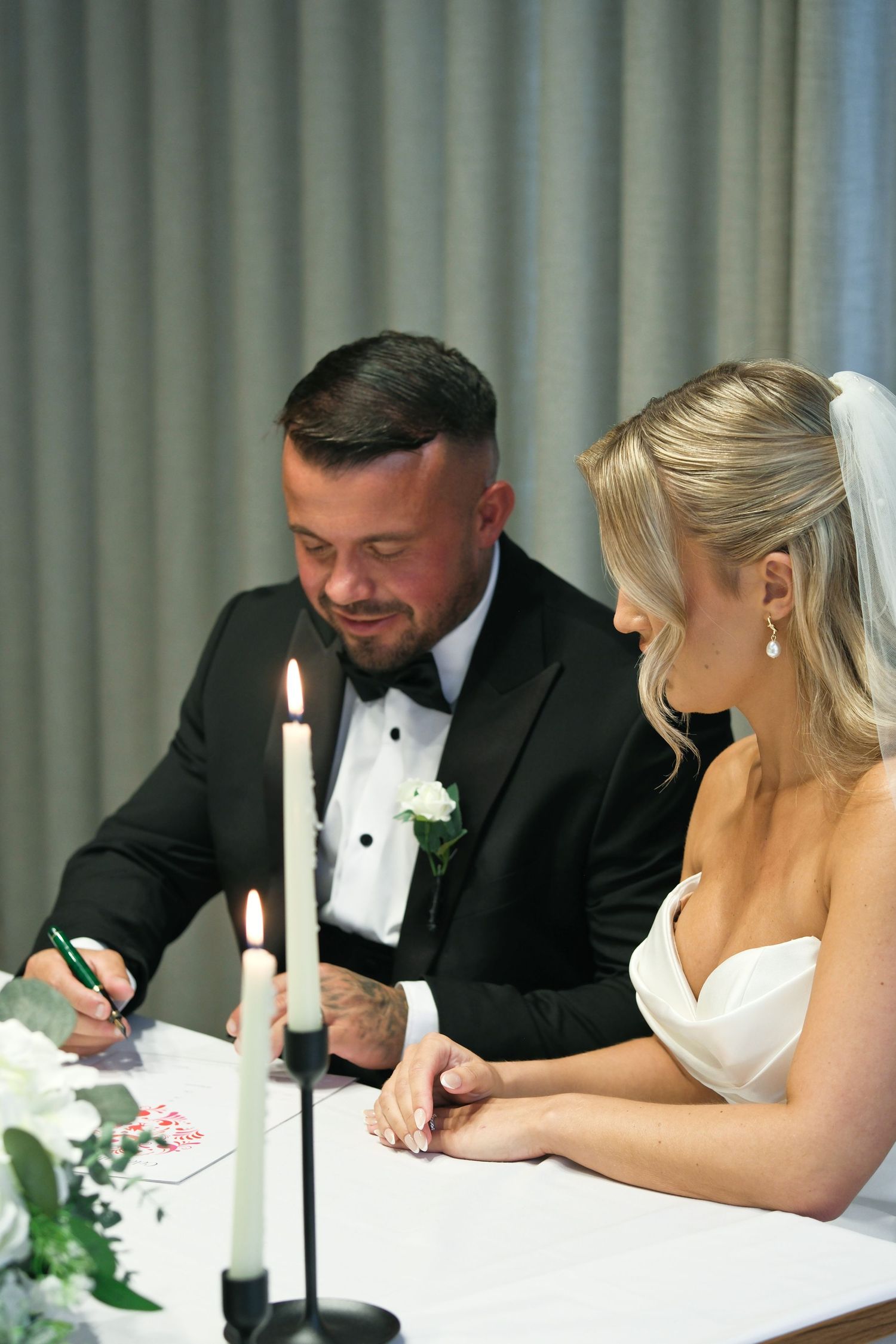 Bride and groom signing wedding registry at candlelit table, groom in black tuxedo with boutonniere, bride in white gown.