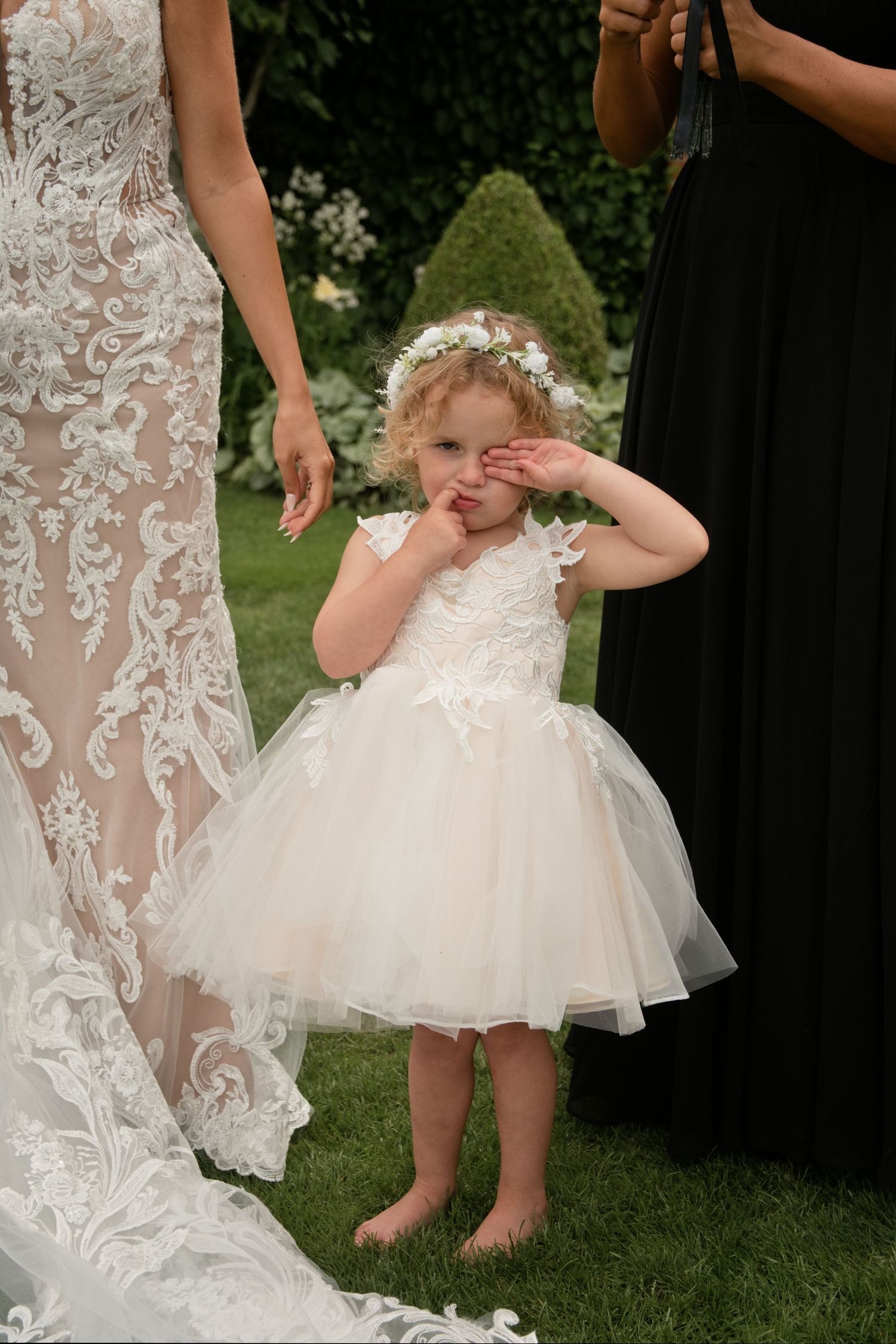 Cute flower girl in white tulle dress with floral crown rubbing her eyes at wedding ceremony.