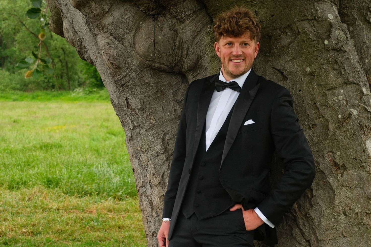 Handsome groom in black tuxedo with bow tie leaning against a large tree outdoors.