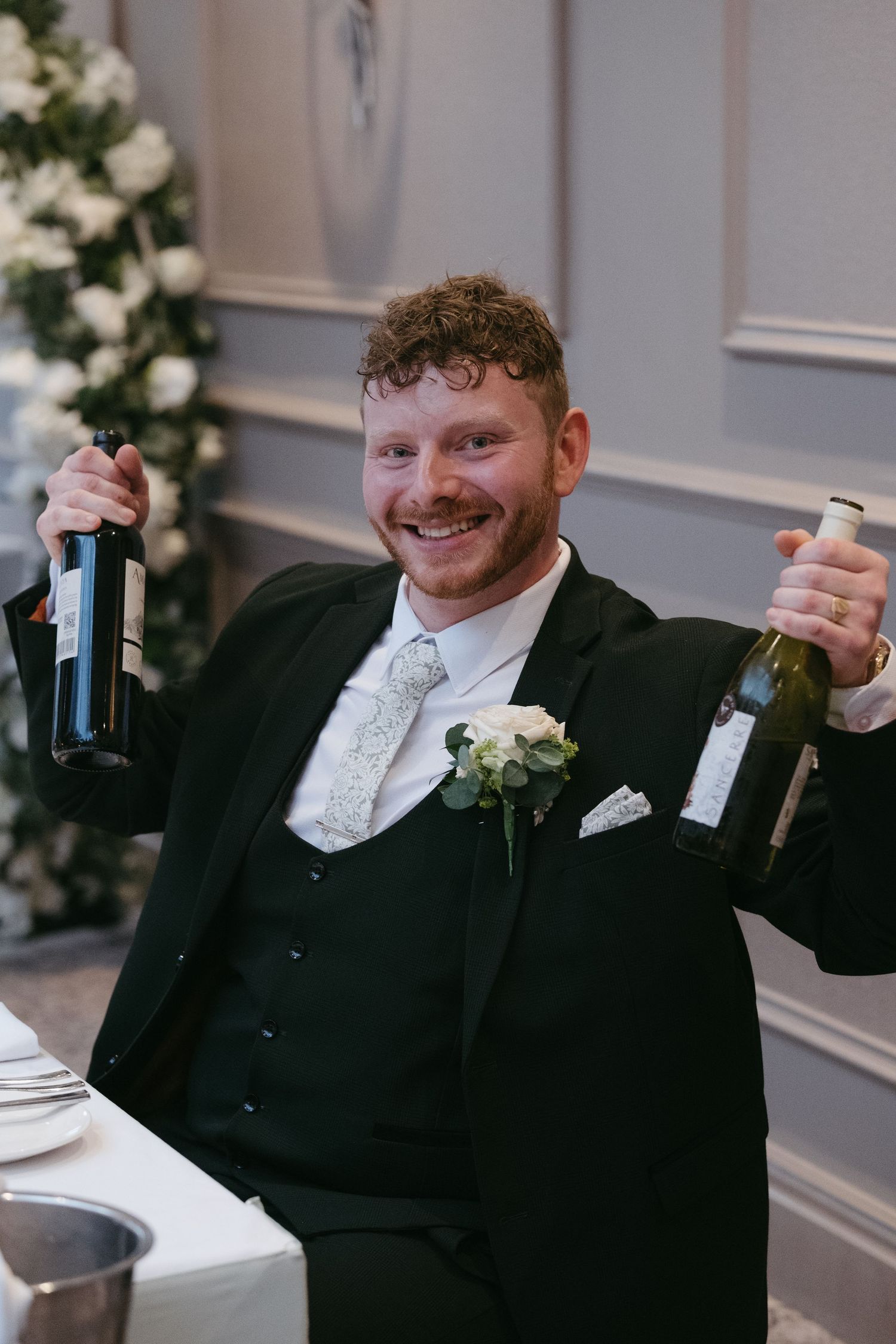 Smiling groom in black suit holds two wine bottles at wedding reception with floral decor behind him.