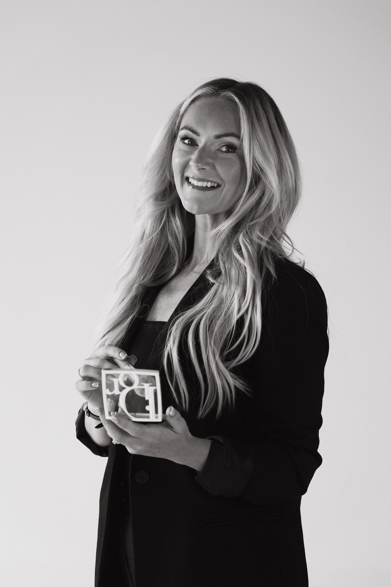 Smiling blonde woman in black blazer holding a small decorative object, black and white studio portrait photo.