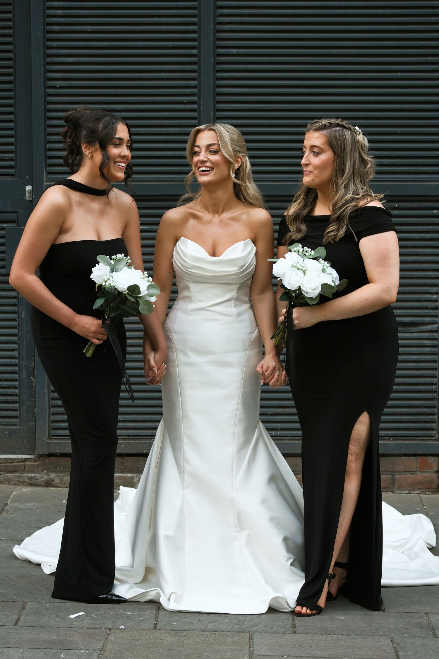 Smiling bride in white mermaid gown poses with two bridesmaids in black dresses holding white rose bouquets.
