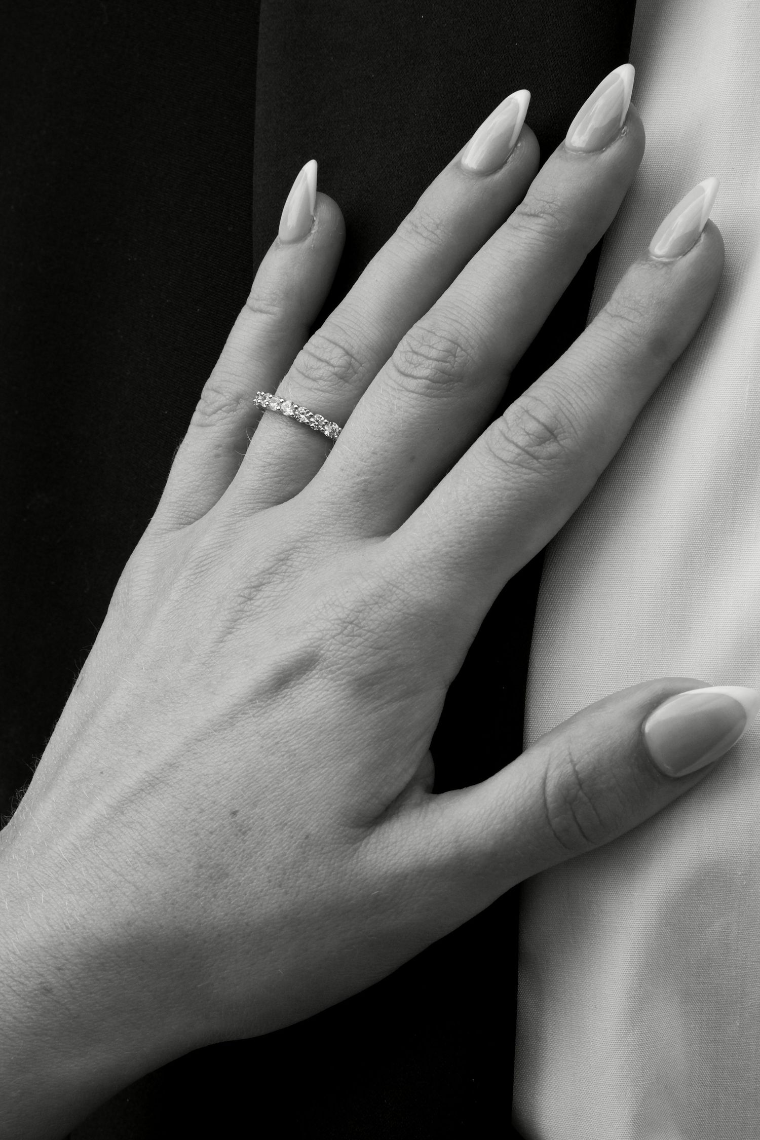 Black and white close-up of bride's hand with diamond eternity band resting on groom's black tuxedo.