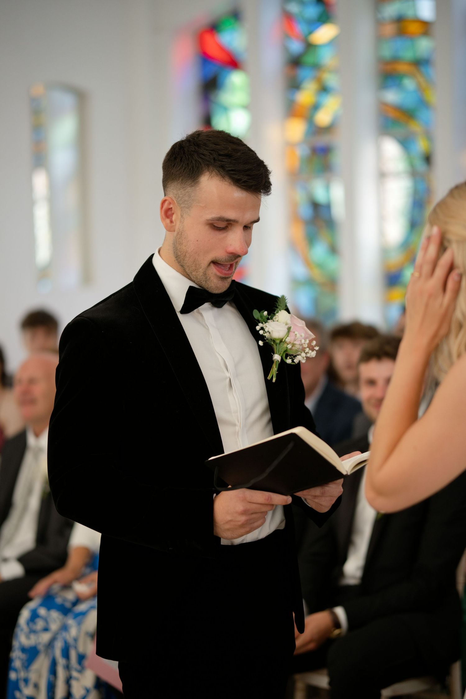 Groom in black tuxedo reading wedding vows from book at church ceremony.