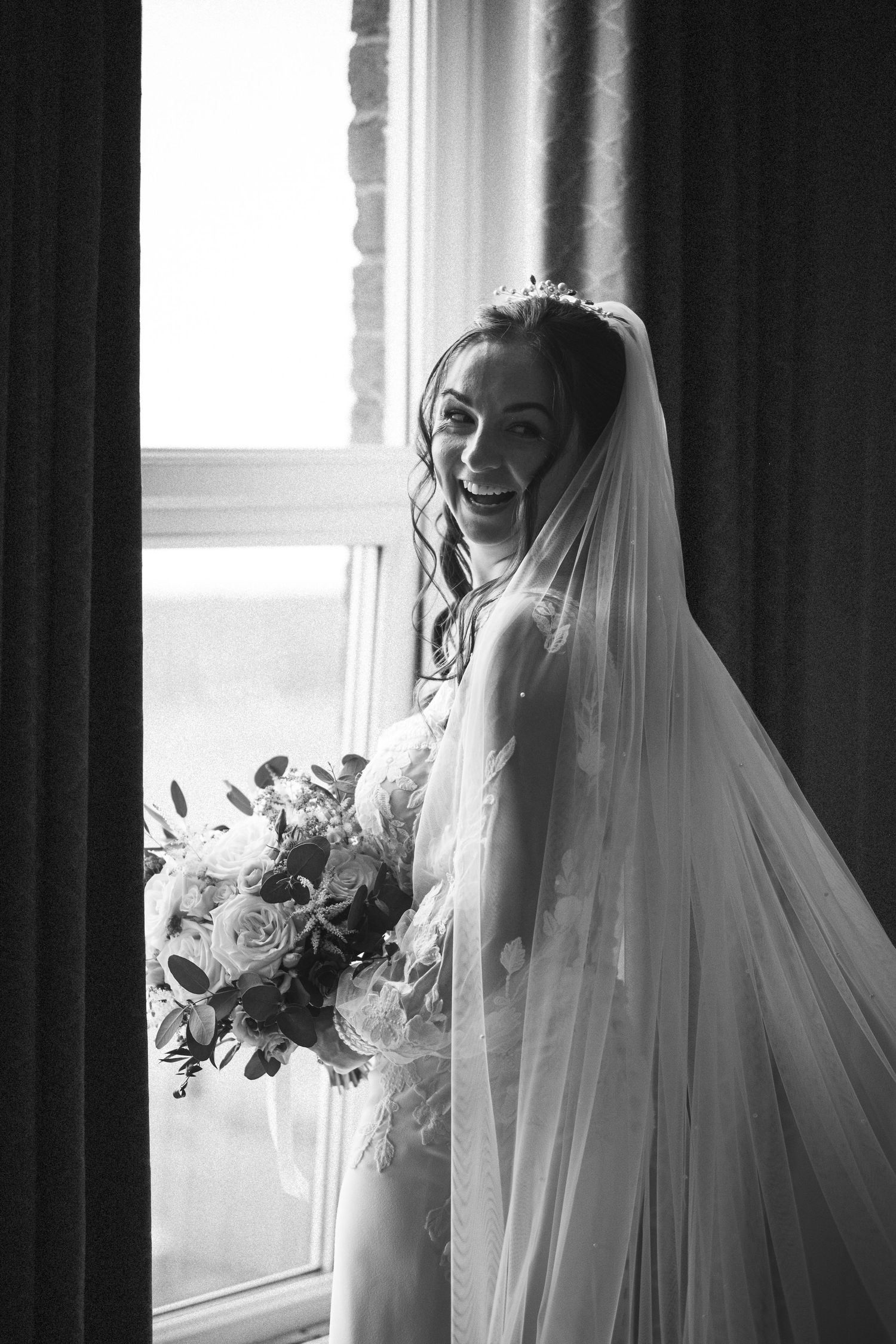 Radiant bride in lace wedding dress and veil holds bouquet while standing by a window in black and white photo.