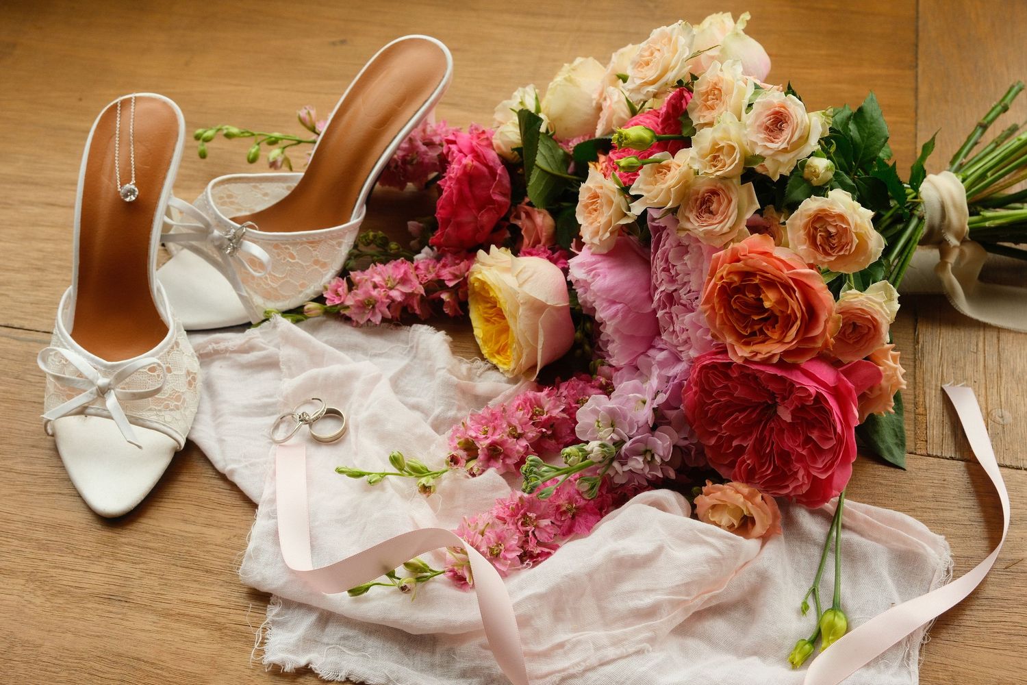 White bridal heels, wedding rings, and a lush bouquet of pink, peach, and coral roses arranged on a wooden floor.