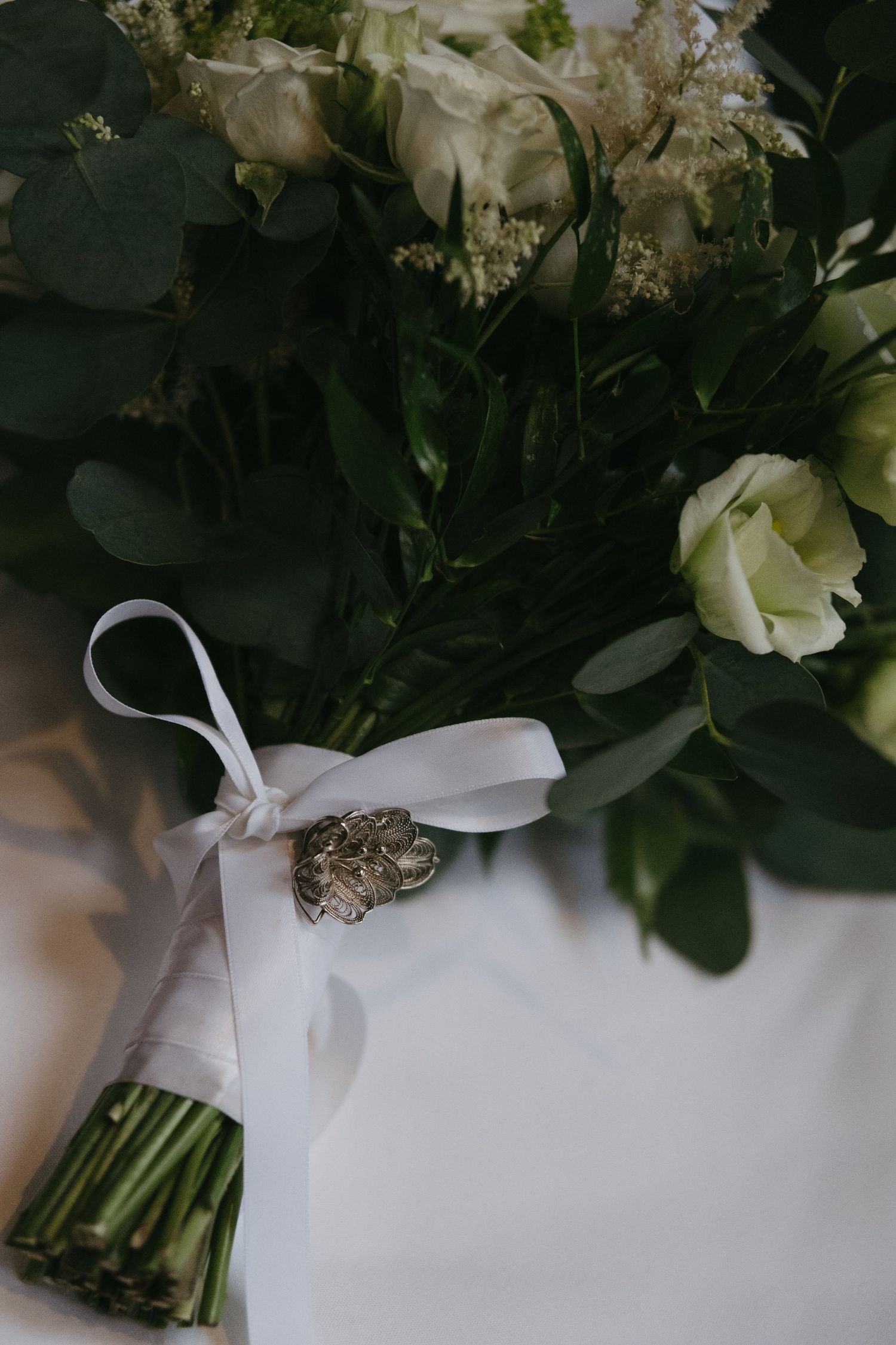 Elegant bridal bouquet of white roses and greenery tied with white ribbon and vintage brooch detail.